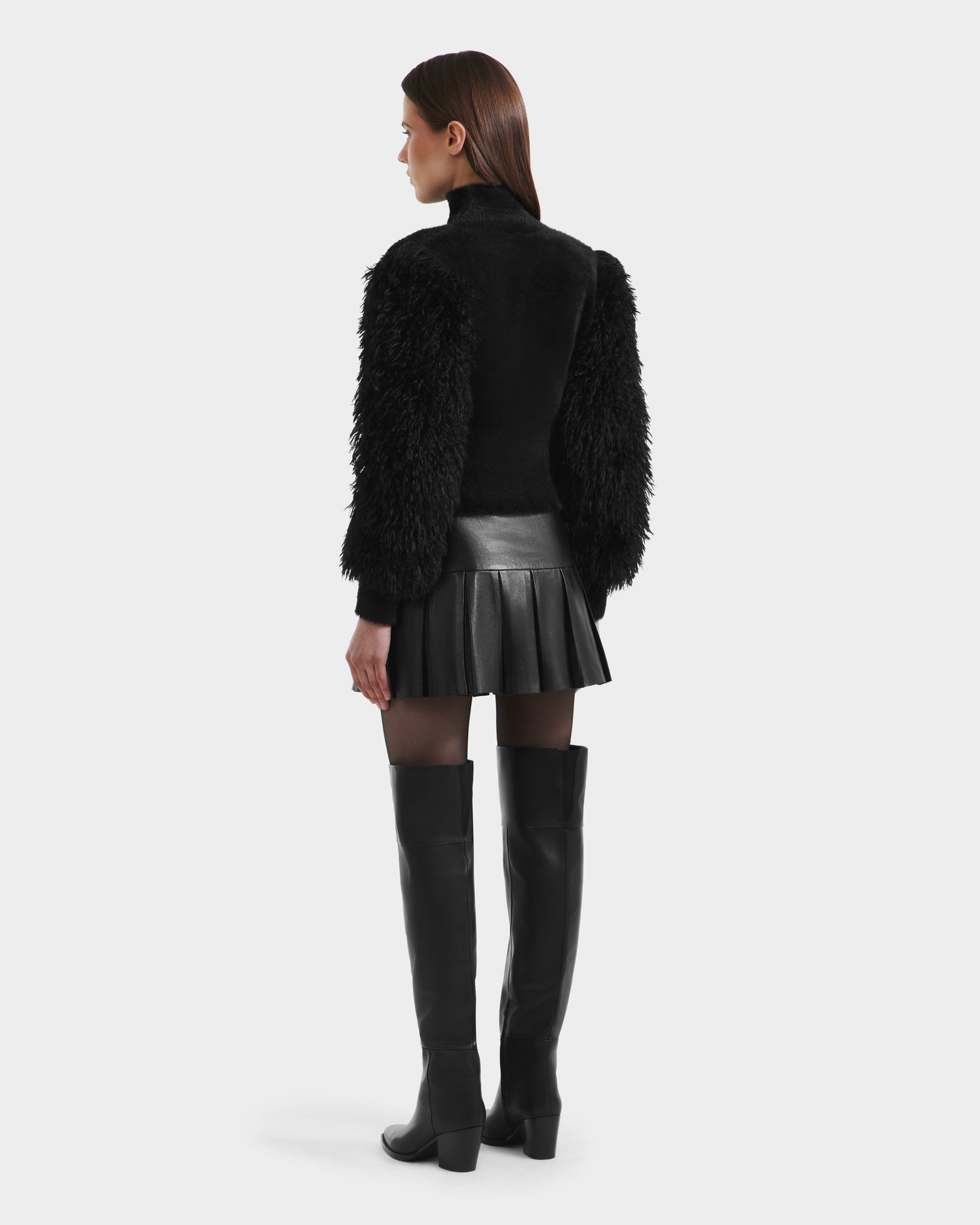 A woman faces away, wearing the Rudsak NOEL - 8525571 Black mock-neck sweater with faux fur sleeves, paired with a black pleated leather skirt, sheer black tights, and tall heeled boots. Perfect for winter layering.