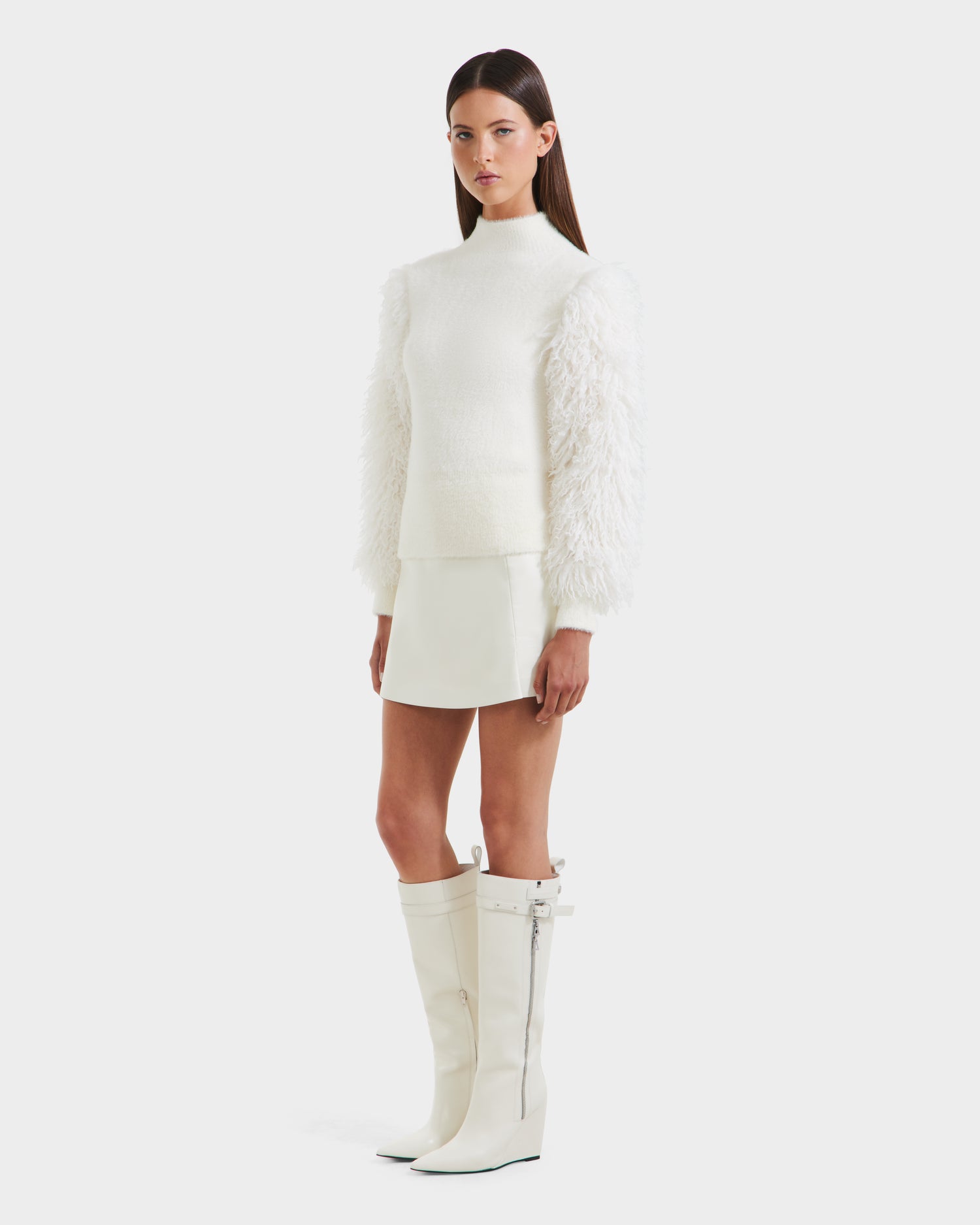A woman poses against a plain white background wearing the Rudsak NOEL - 8525571 Vanilla textured women's mock-neck sweater with faux fur sleeves, paired with a white mini skirt and tall white heeled boots.