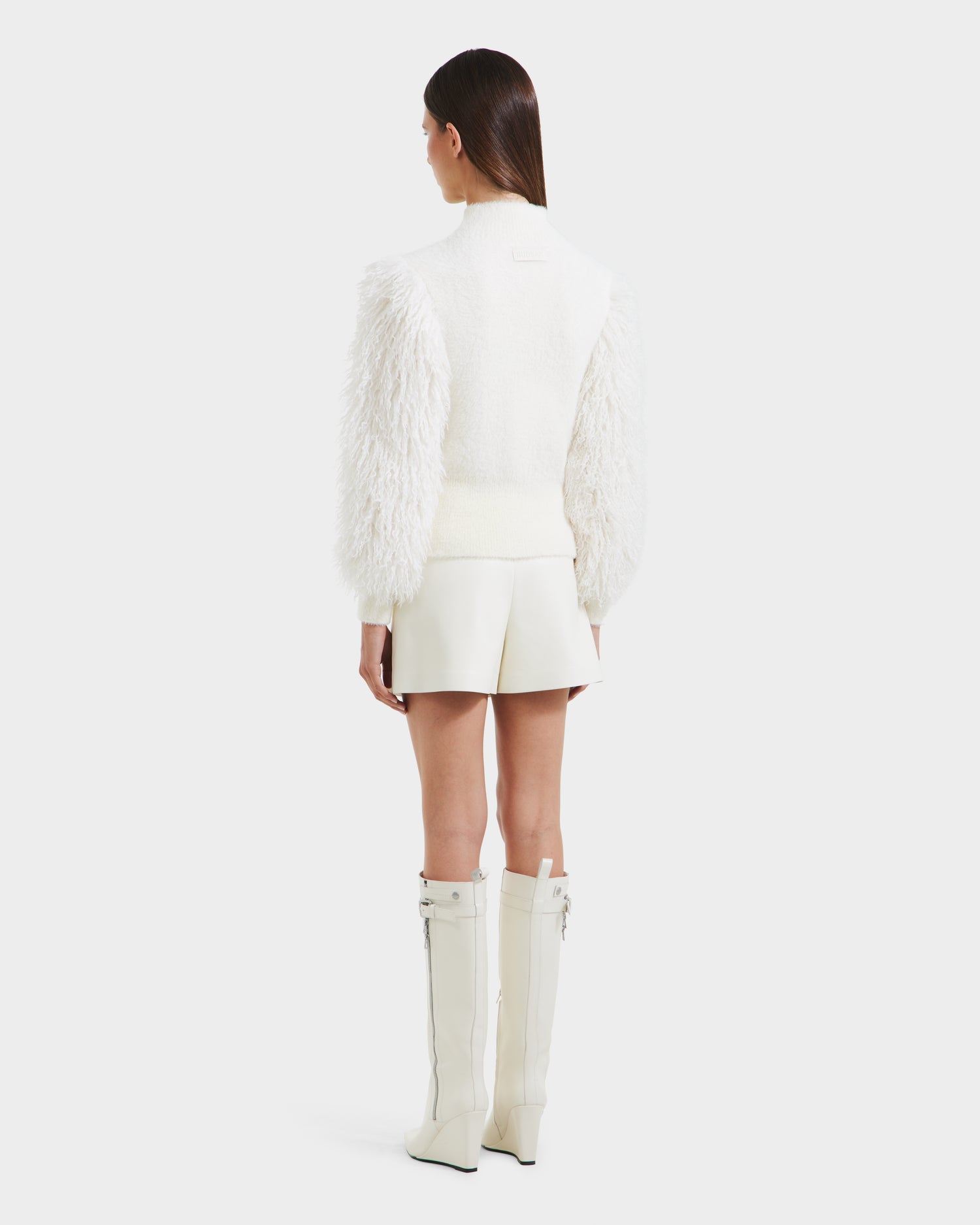 A woman faces away, wearing the Rudsak NOEL - 8525571 Vanilla jacket with faux fur sleeves, a short white skirt, and knee-high white boots. Her straight brown hair is down against a plain white background—perfect winter style.