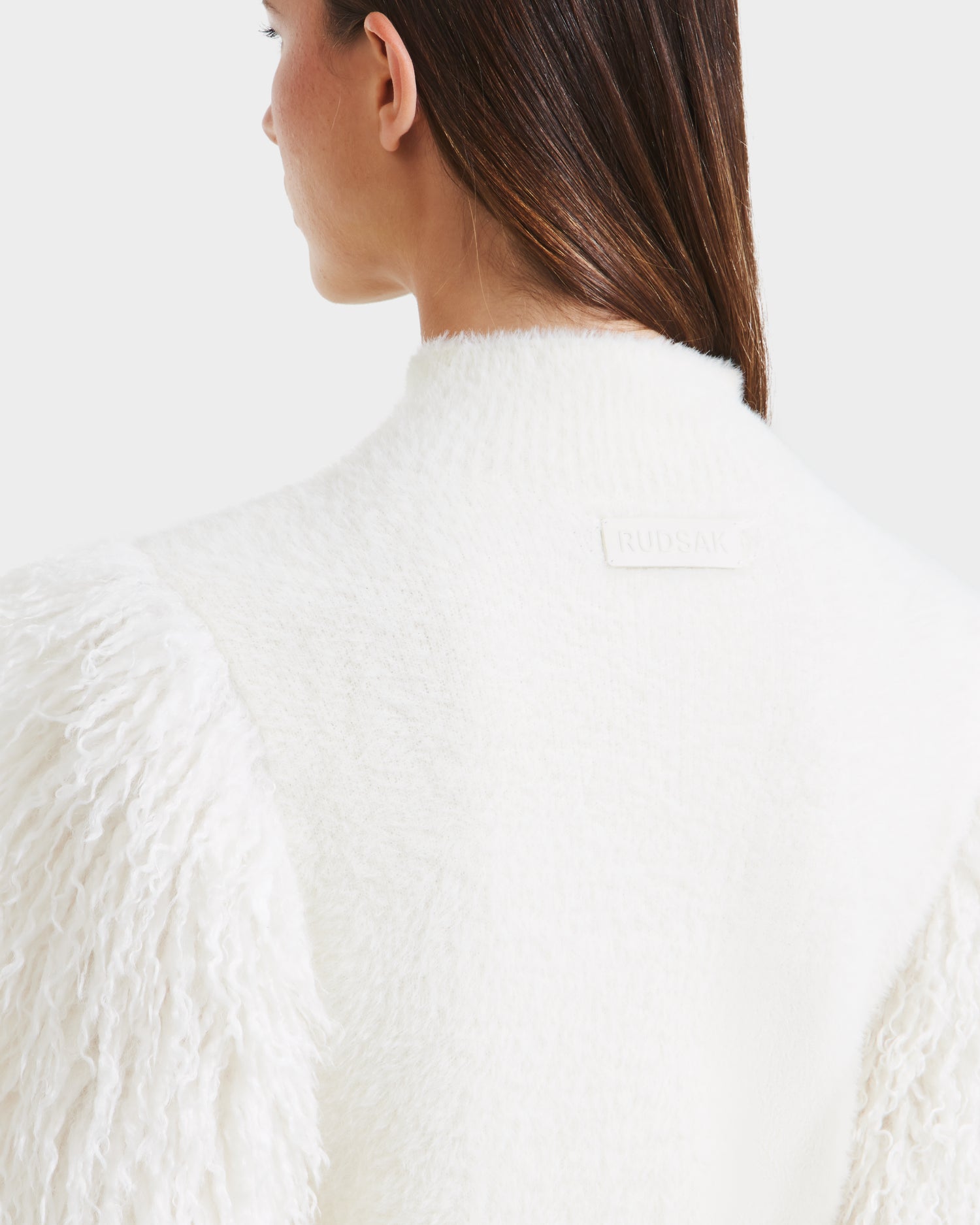 A woman with straight brown hair, seen from the back, wears the Rudsak NOEL - 8525571 Vanilla coat, a white textured winter piece with faux fur sleeves and a small white Rudsak label near the collar.