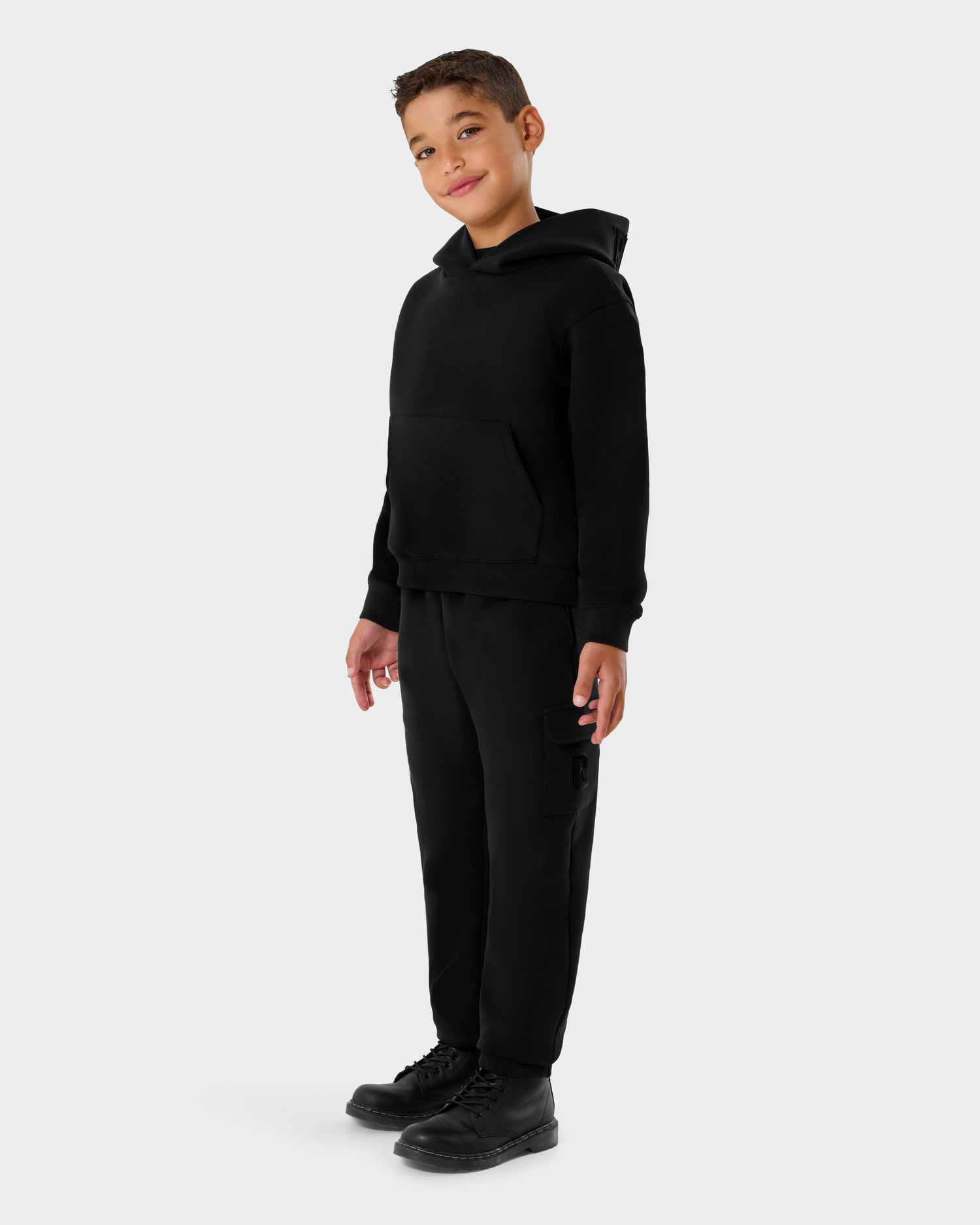 A young boy smiles in front of a light gray background, wearing the Rudsak NOOR - 1525004 Black hoodie, matching black sweatpants, and black boots.