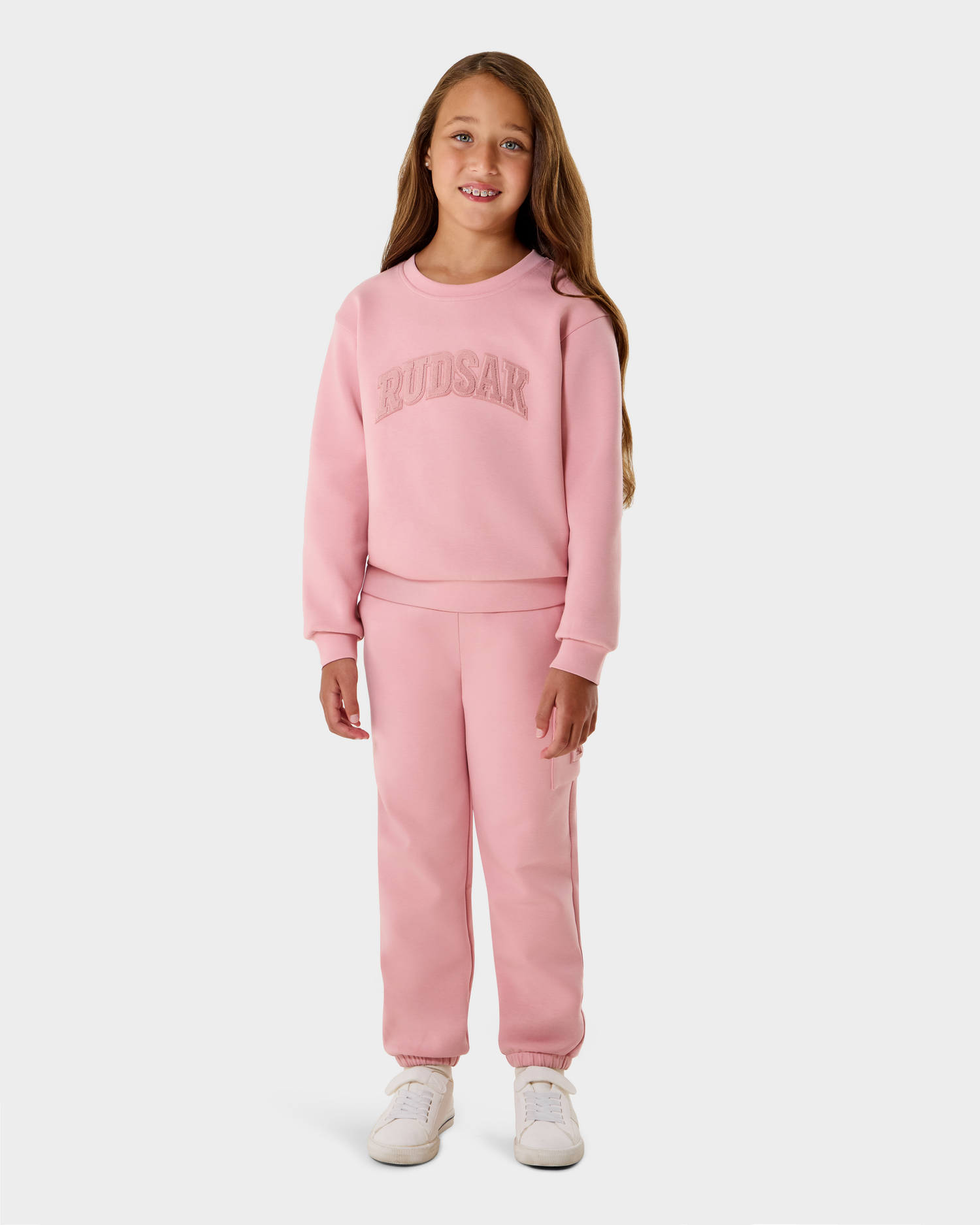 A young girl with long brown hair stands smiling in a matching light pink Rudsak NOOR - 1525004 sweatshirt and sweatpants, white sneakers, and a sweatshirt featuring the brand name. The background is plain white.