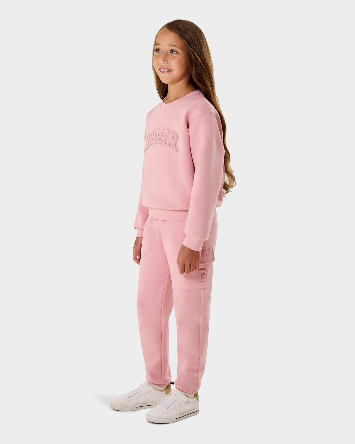 A young girl with long brown hair wears the Rudsak NOOR - 1525004 Light Pink matching sweatshirt and joggers set with white sneakers, standing and smiling against a plain white background.