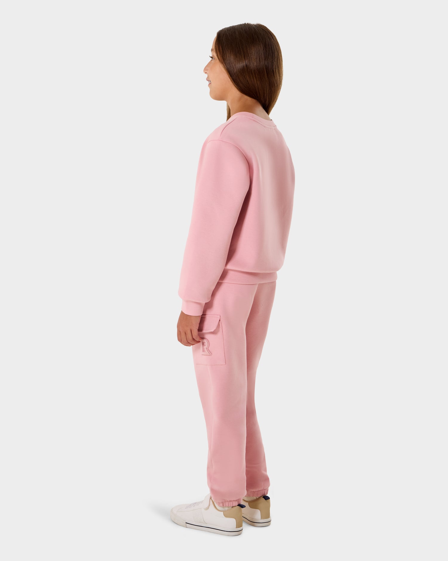 A young girl stands sideways against a white background, wearing the Rudsak NOOR - 1525004 Light Pink sweatshirt and matching sweatpants with white sneakers.