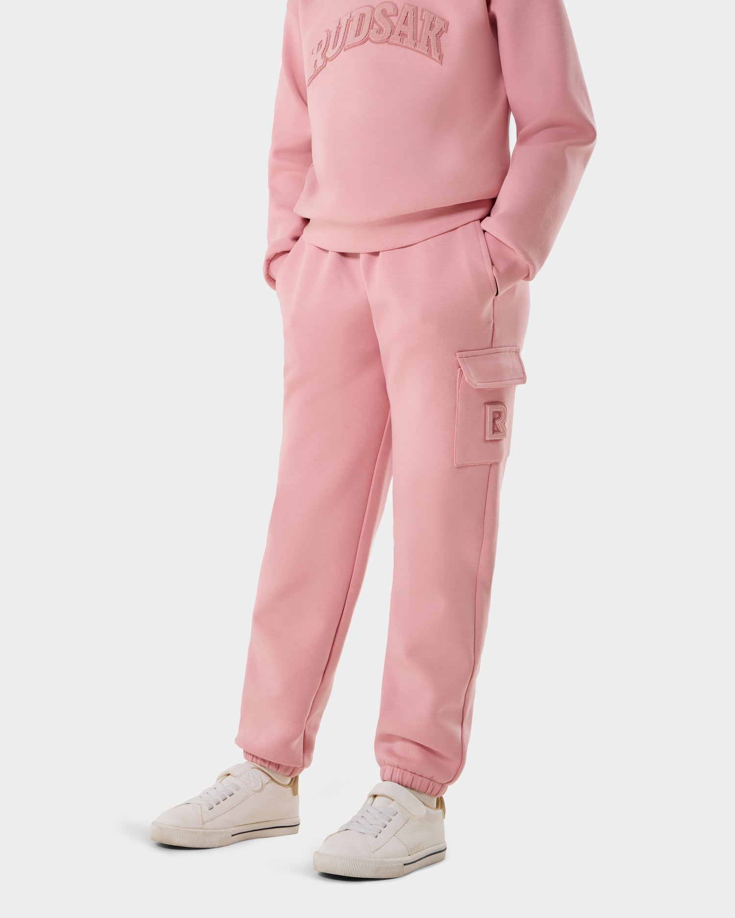 A person wears the Rudsak NOOR - 1525004 Light Pink sweatshirt and matching cargo sweatpants with white sneakers, hands in pockets, showing only the lower half of the body against a plain background.