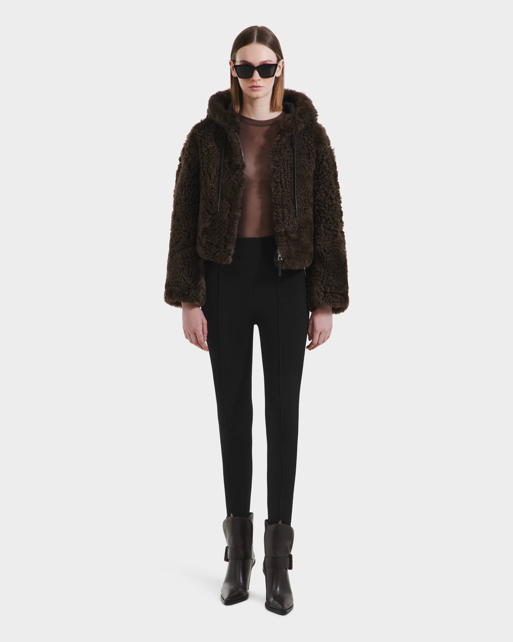A woman faces forward wearing the Rudsak NORAH - 8125506 Black/Espresso reversible shearling leather jacket, a sheer brown top, black high-waisted pants, cuffed pointed ankle boots, and black sunglasses against a white background.