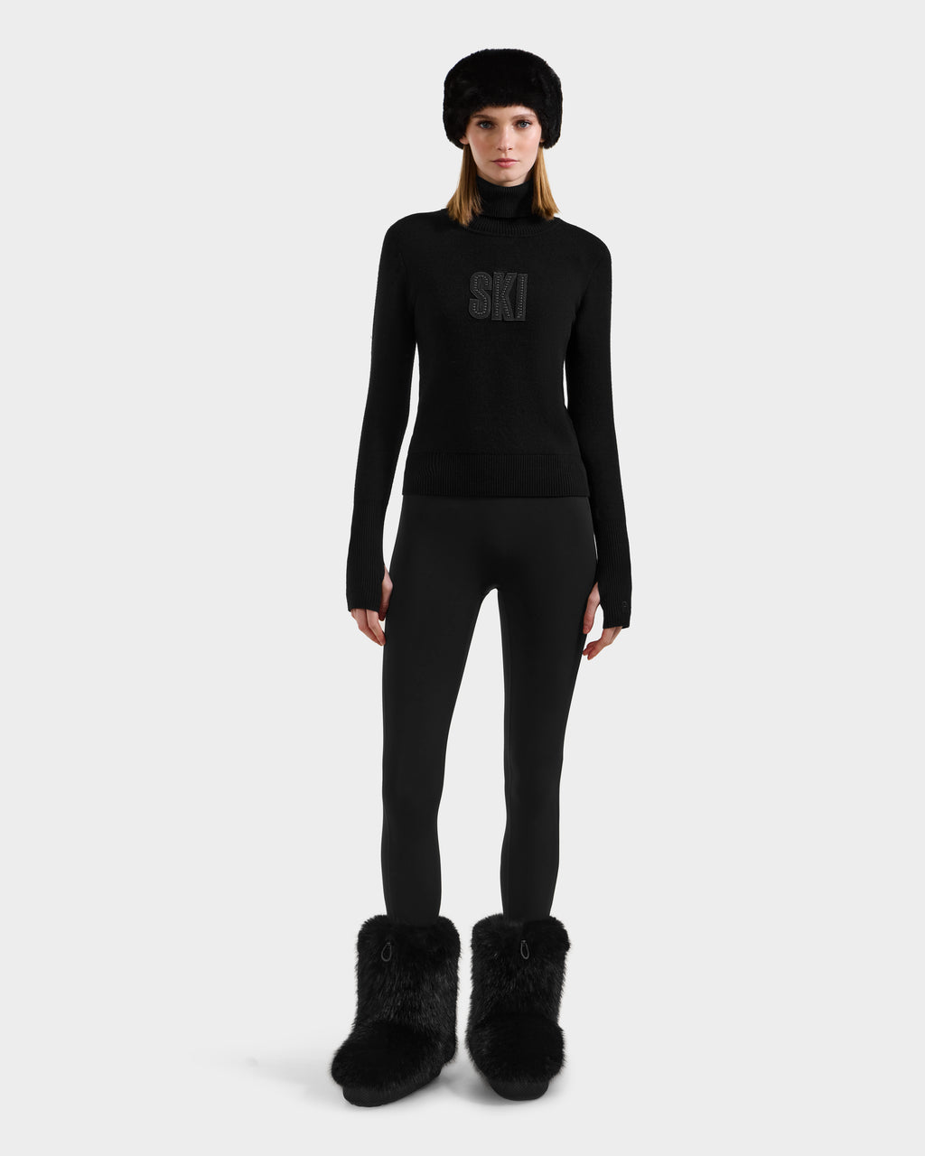 A woman faces forward against a plain light background, wearing the Rudsak NOVIE - 8525659 Black après-ski sweater with SKI on the front, paired with black leggings, a black fur hat, and large black fur boots.