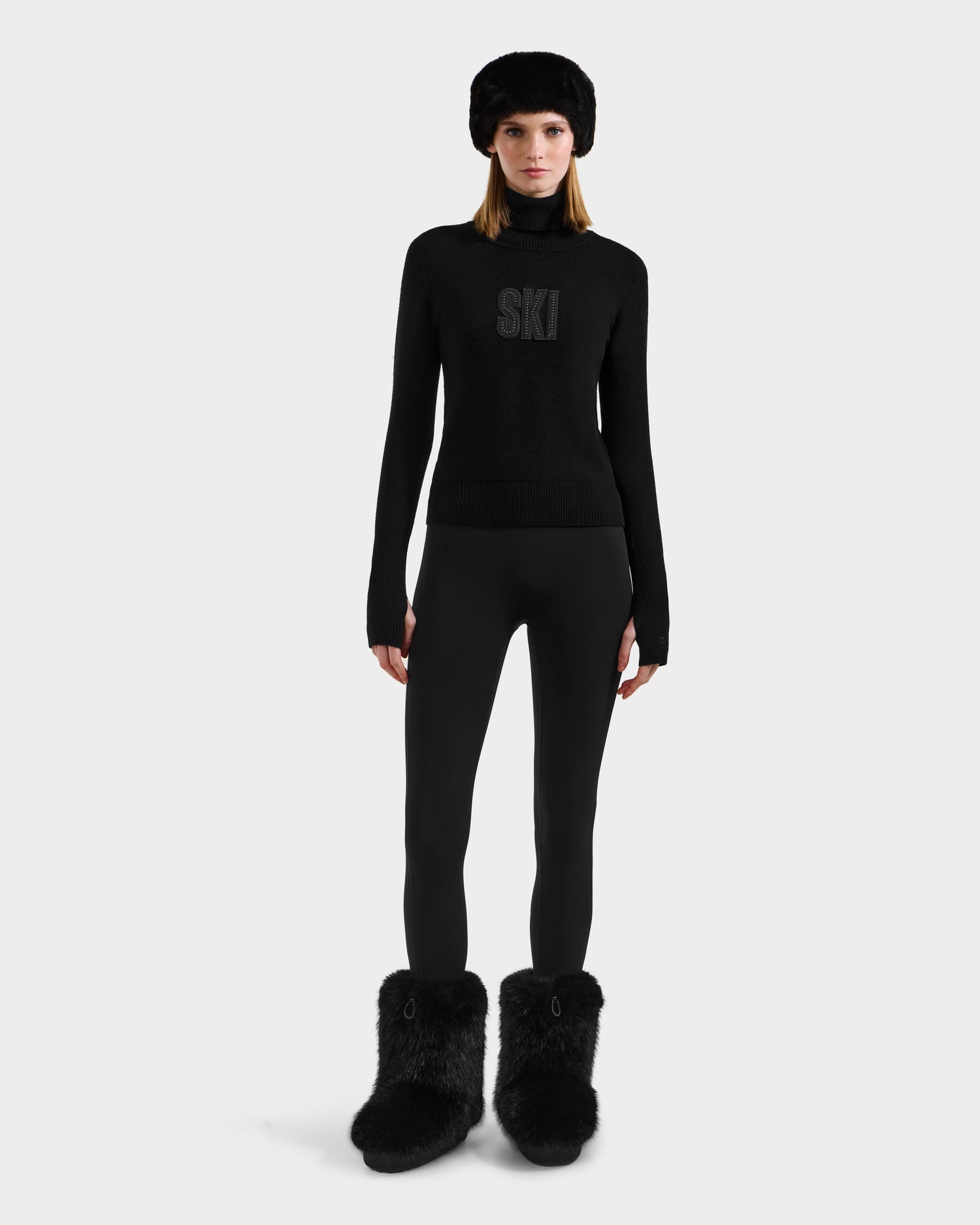 A woman faces forward against a plain light background, wearing the Rudsak NOVIE - 8525659 Black après-ski sweater with SKI on the front, paired with black leggings, a black fur hat, and large black fur boots.
