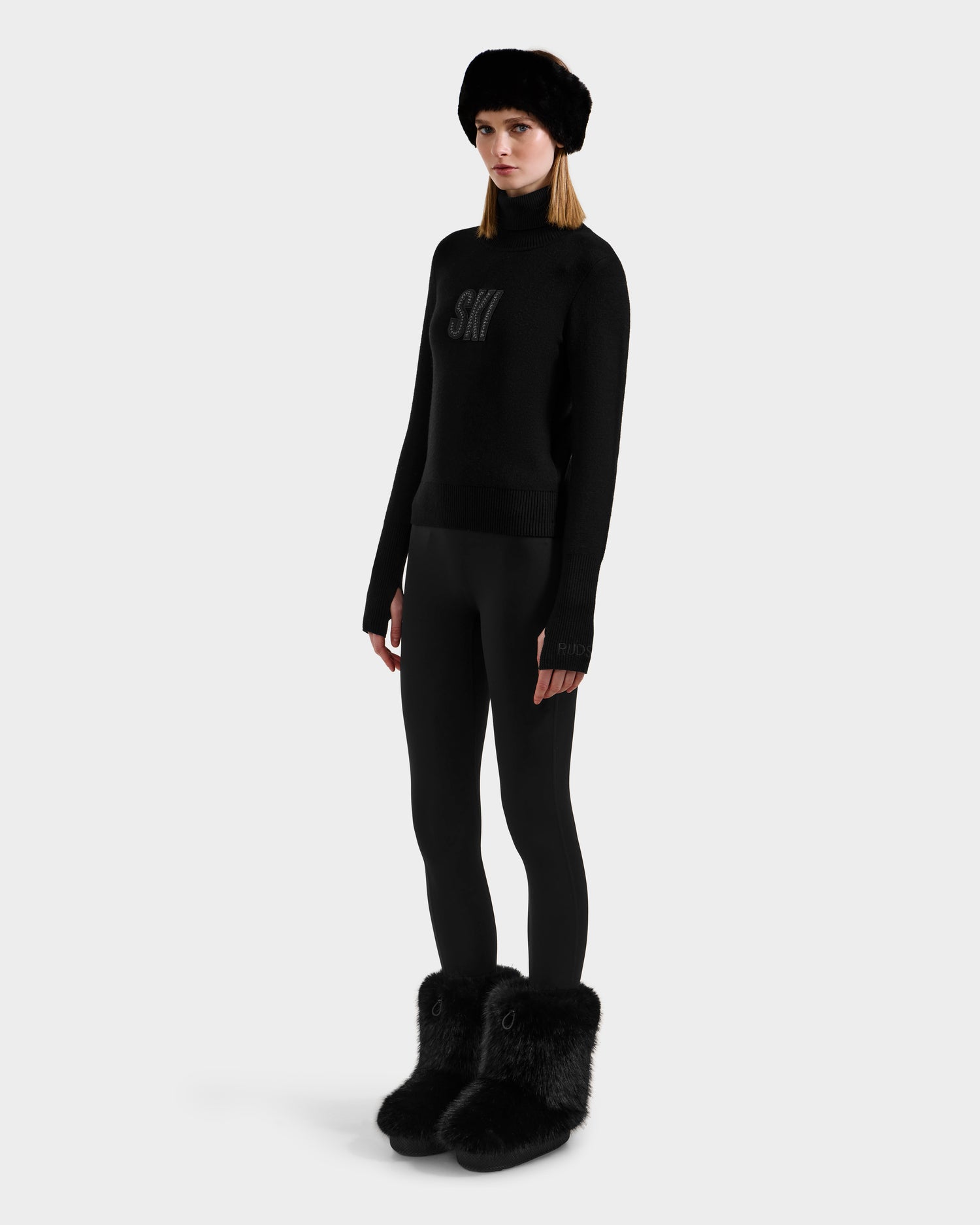 A woman wears the Rudsak NOVIE - 8525659 Black, an all-black après-ski outfit with a fur hat, SKI sweater, fitted pants, and furry boots, standing against a plain white background.
