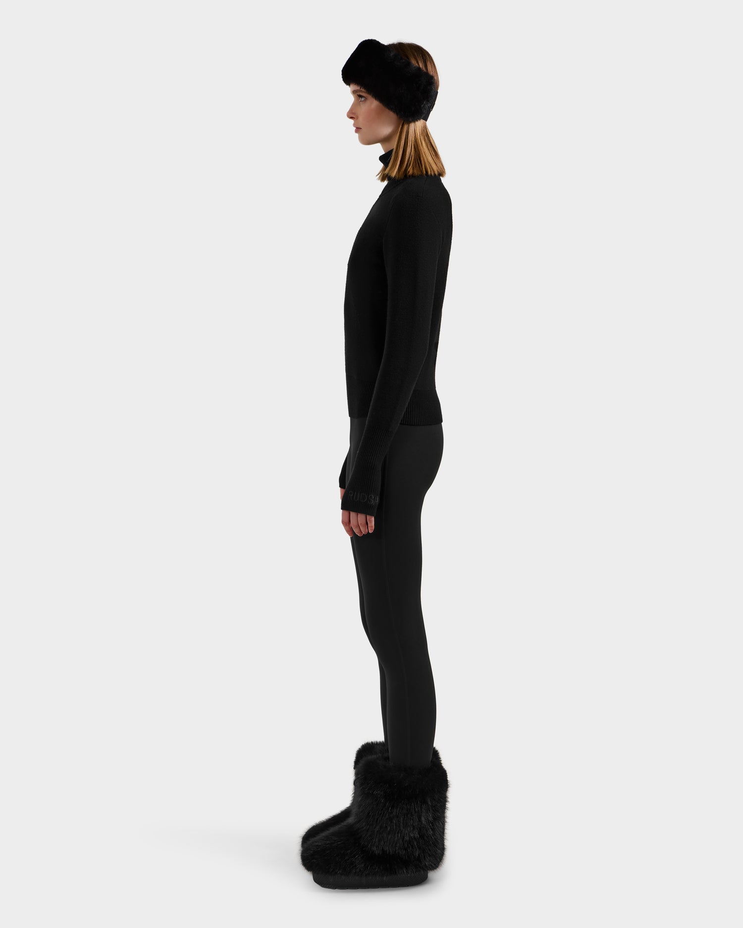 A woman stands in profile against a plain background, wearing Rudsak’s NOVIE - 8525659 Black knit turtleneck, leggings, a black fur headband, and oversized black fur boots.