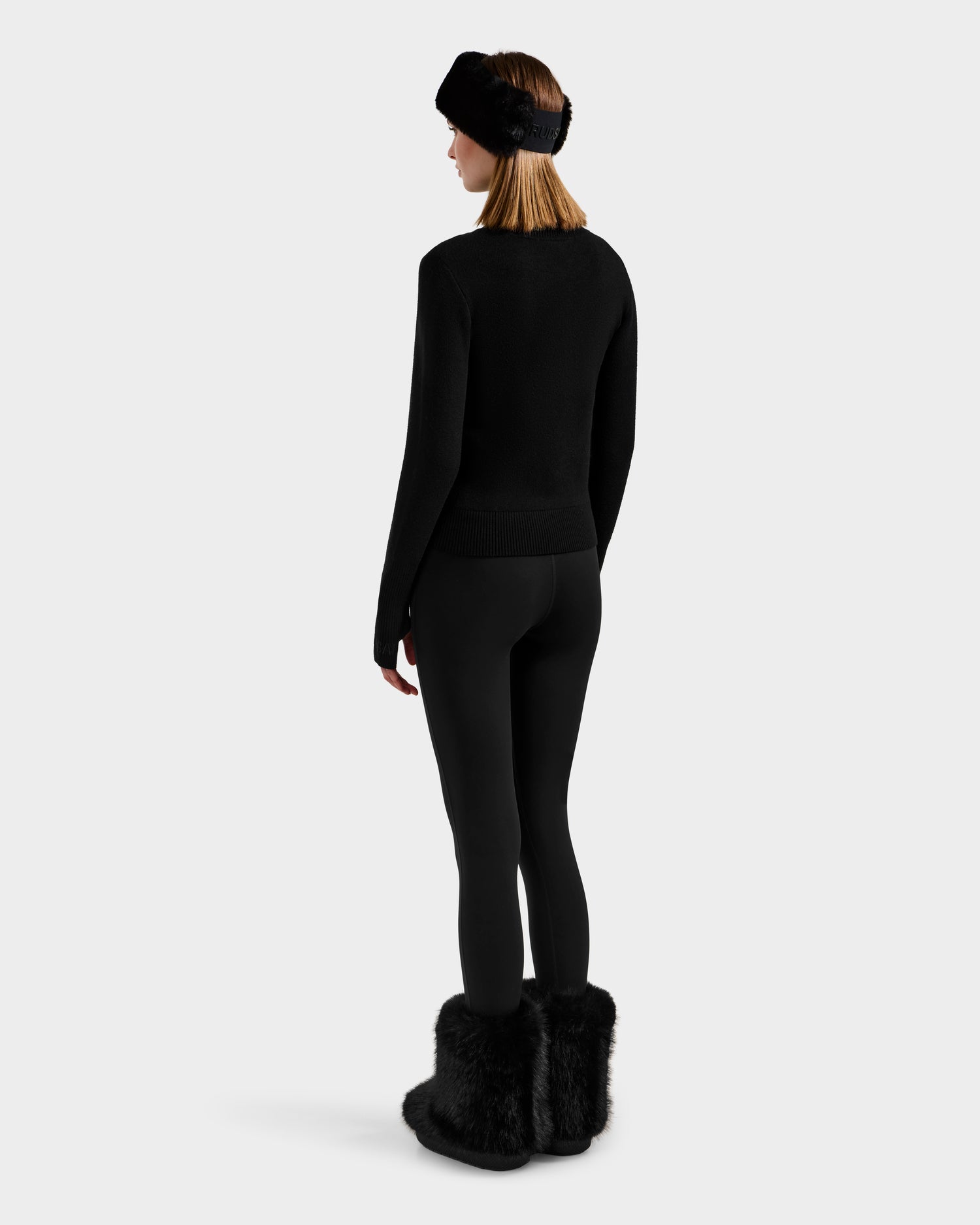 A woman stands facing away, wearing the Rudsak NOVIE - 8525659 Black women's knit turtleneck, black leggings, black furry boots, and a black furry headband against a plain white background.
