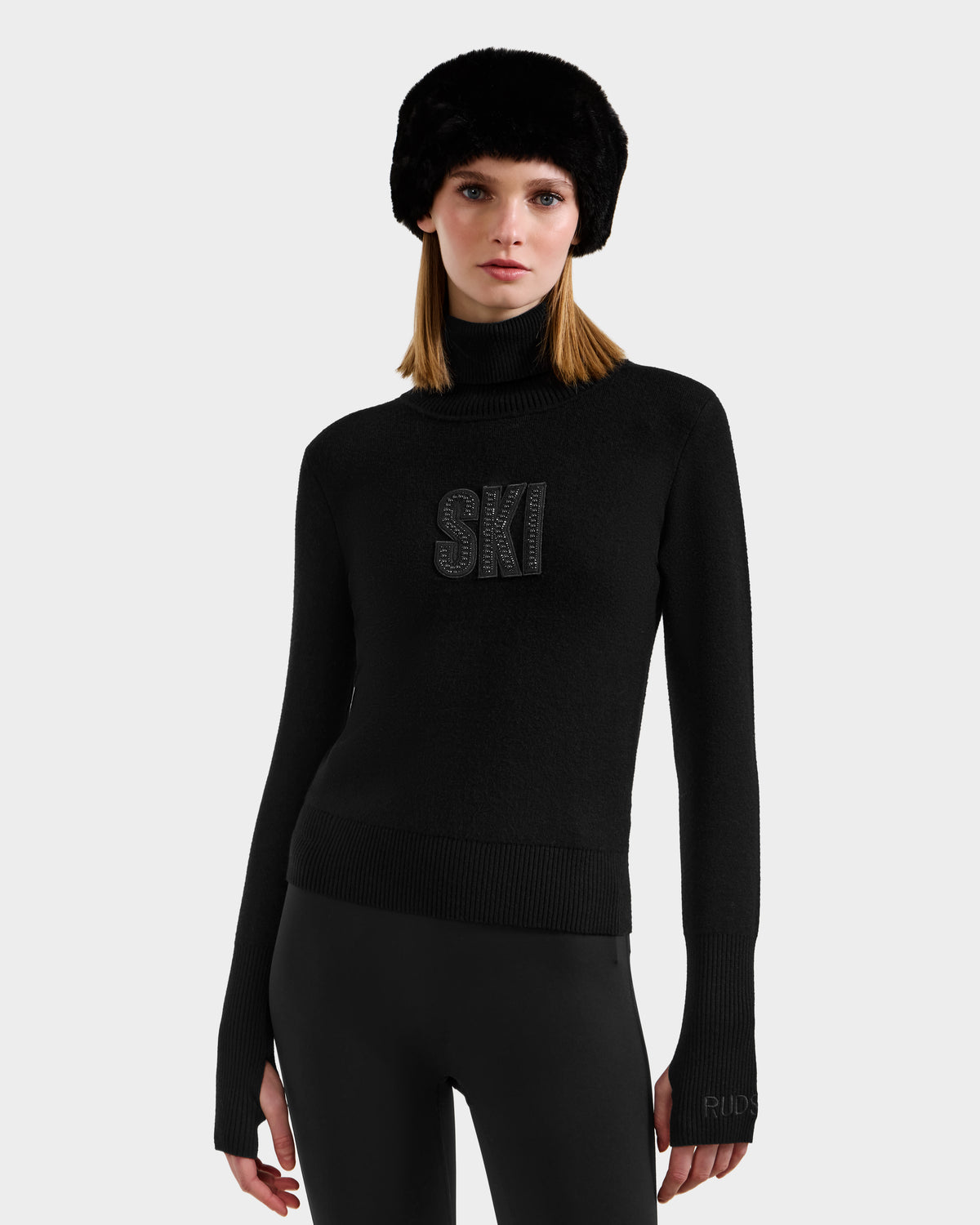 A woman models the Rudsak NOVIE - 8525659 Black ski sweater with "SKI" on the front, paired with black fitted pants and a furry hat, creating an effortlessly chic après-ski look against a light gray background.