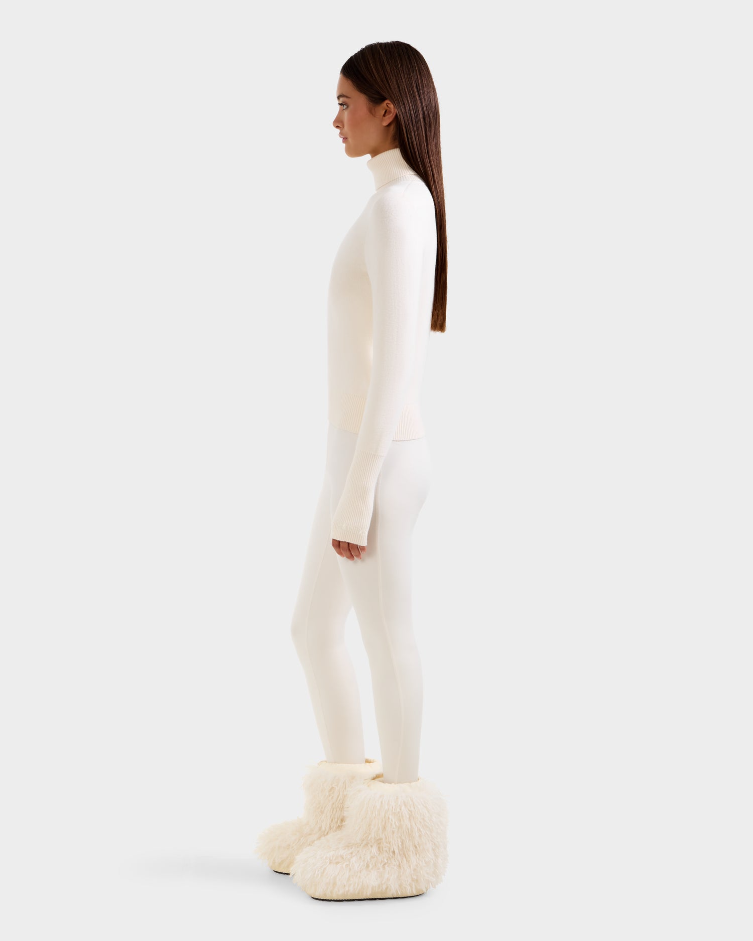 A woman in profile wears the Rudsak NOVIE - 8525659 Vanilla boots, paired with a fitted knit turtleneck and white leggings. Her straight brown hair falls past her shoulders against a plain white background.