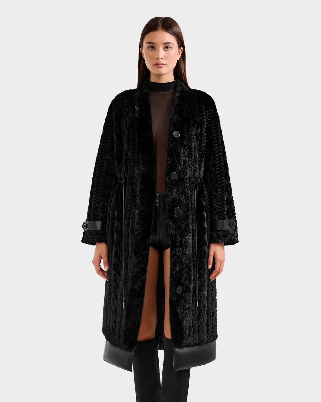 A woman stands facing forward in the Rudsak ORIANA - 8125545 Black, a long textured faux fur coat with vegan leather trims, layered over a sheer black top and black shorts, set against a plain light gray background.