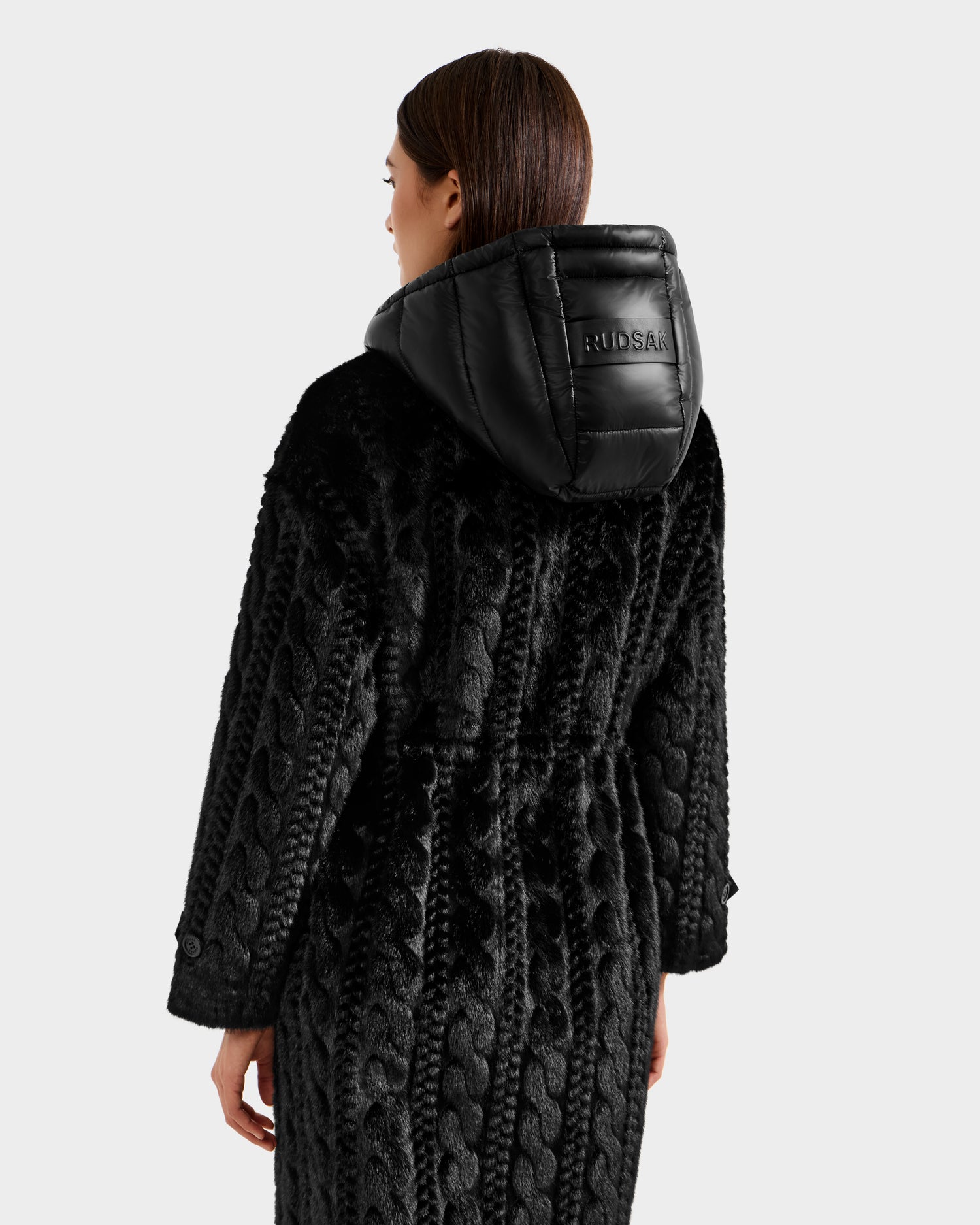 A person with straight brown hair wears the Rudsak ORIANA - 8125545 Black textured coat with vegan leather trims and a shiny hood embossed with RUDSAK on the back, standing against a plain light background.