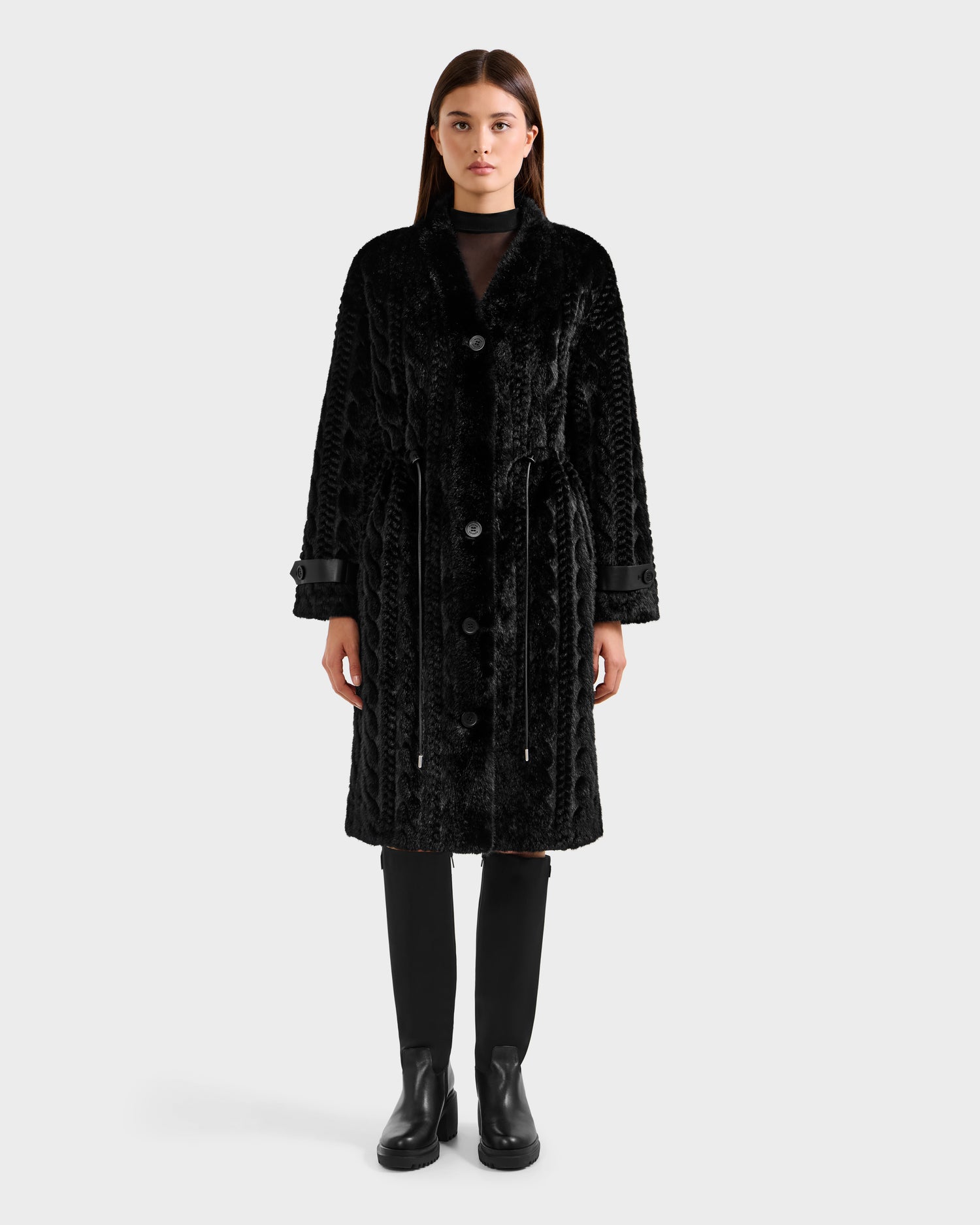 A woman with straight brown hair and a neutral expression wears the Rudsak ORIANA - 8125545 Black women’s faux fur coat with vegan leather trims and large buttons, paired with knee-high black boots, standing against a plain light background.
