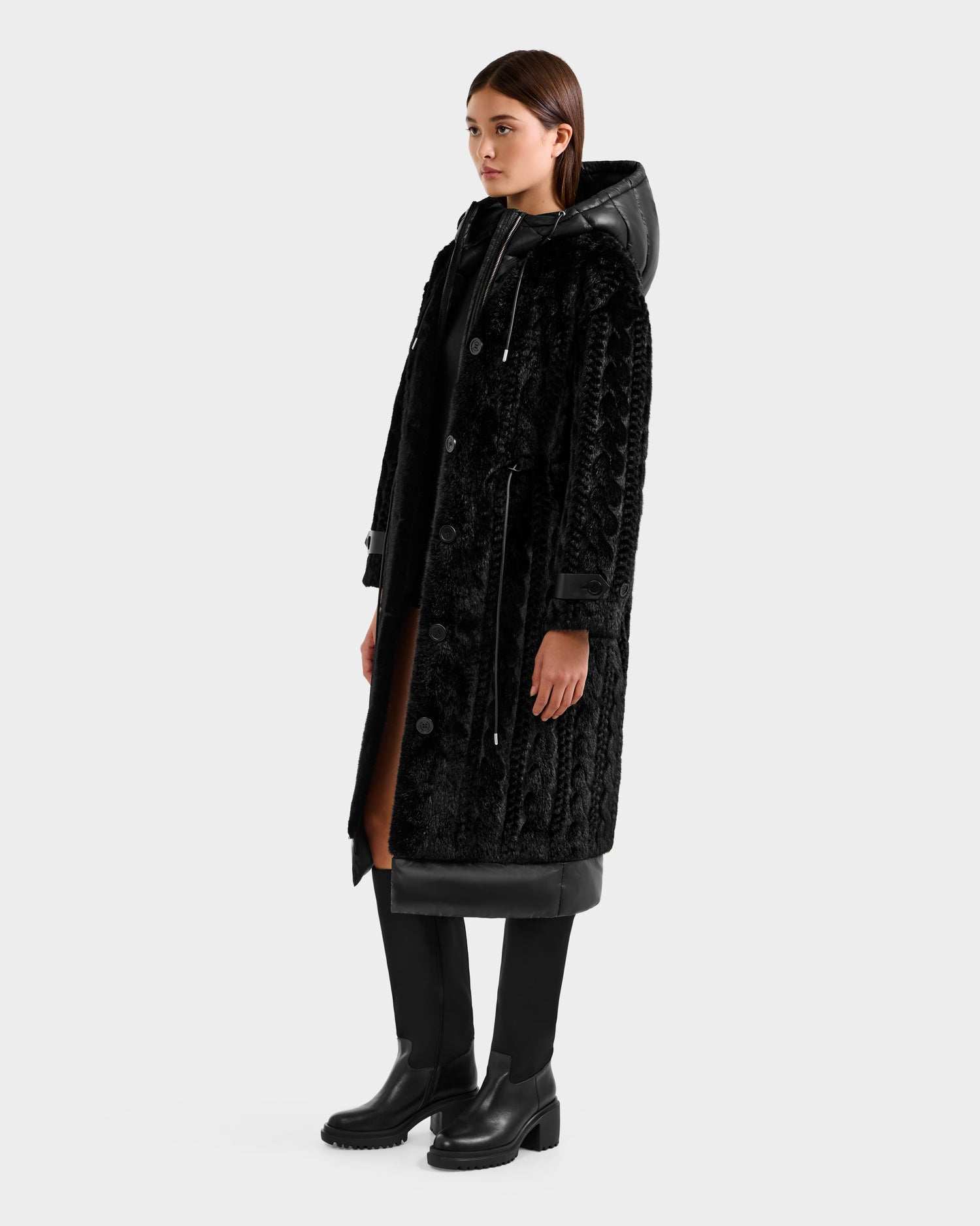 A woman stands facing slightly right, wearing the Rudsak ORIANA - 8125545 Black coat with vegan leather trims, black pants, and chunky boots. Her hands rest at her sides against a plain light background.