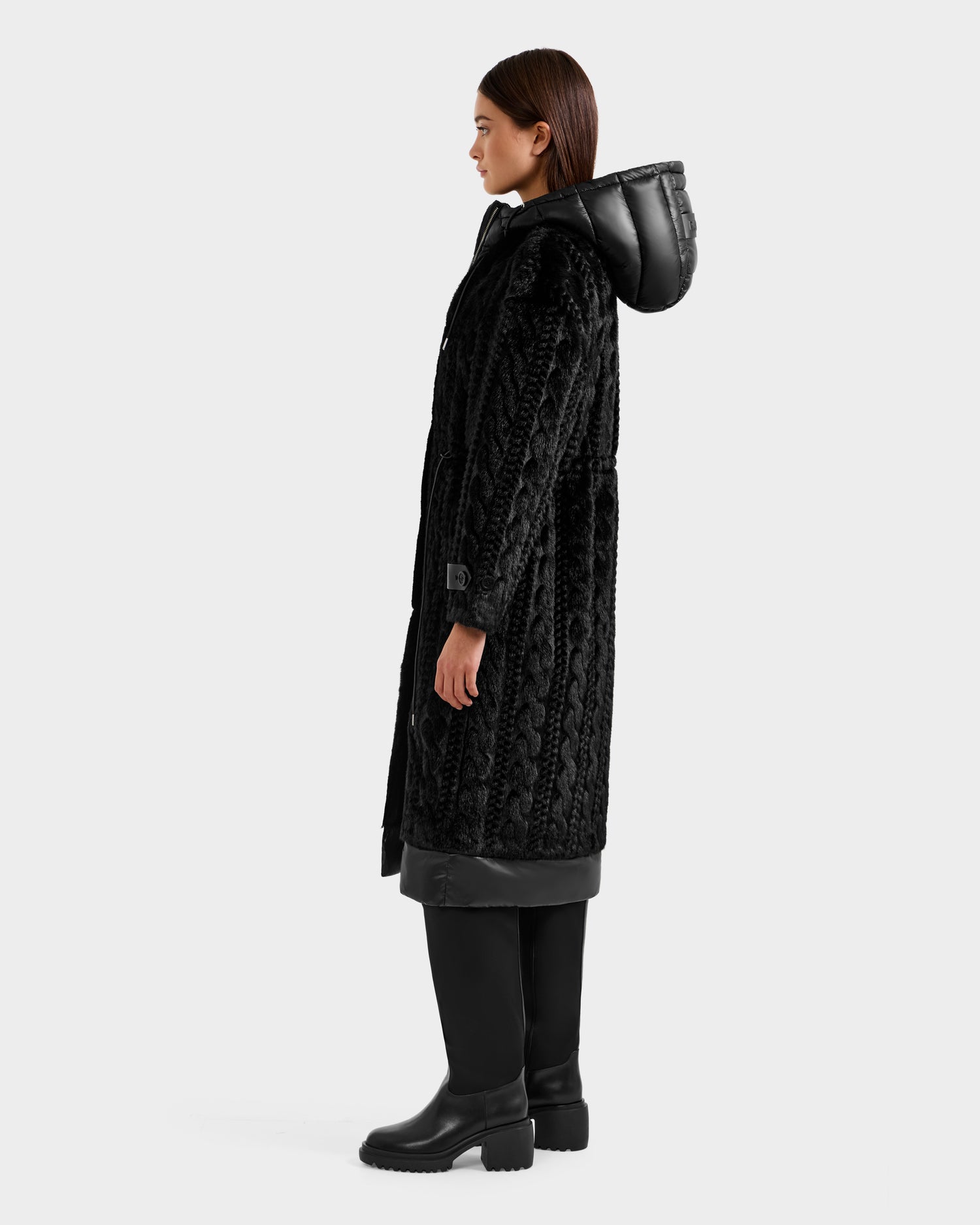A woman stands sideways against a plain light background, wearing the Rudsak ORIANA - 8125545 Black textured coat with vegan leather trims and a hood, paired with black pants and chunky boots.