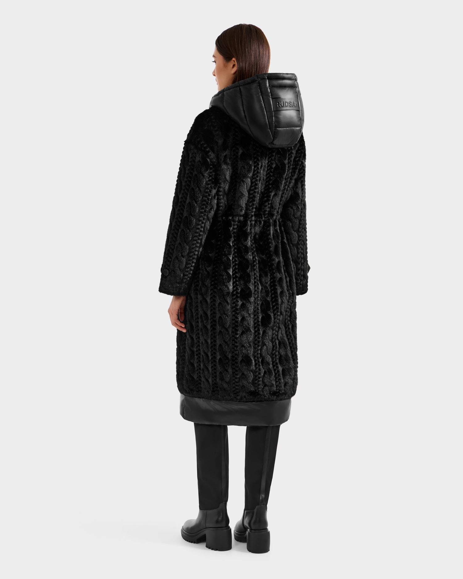 A woman faces away, wearing Rudsak's ORIANA - 8125545 Black textured coat with vegan leather trims and a hood, paired with black knee-high boots against a plain white background.