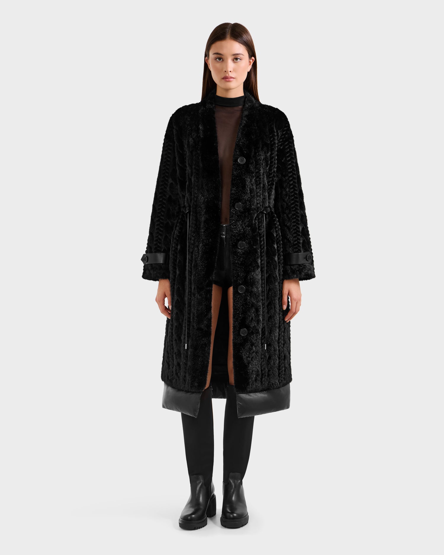 A woman faces forward, wearing the Rudsak ORIANA - 8125545 Black long textured coat with vegan leather trim, black boots, and a sheer black top over shorts, set against a plain light background.