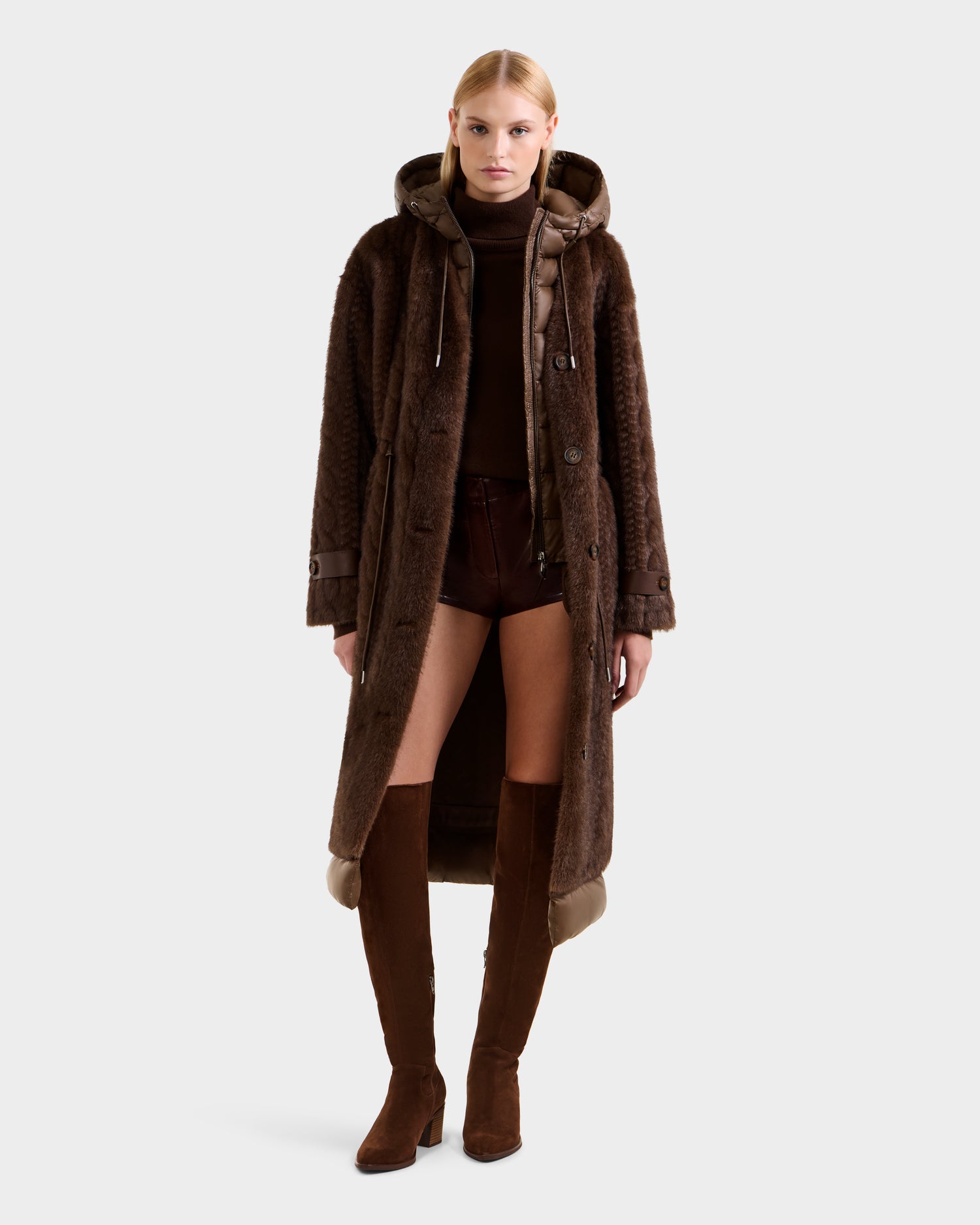 A woman stands facing forward in the Rudsak ORIANA - 8125545 Mocha faux fur coat with vegan leather trim, styled with a brown turtleneck, brown shorts, and knee-high brown suede boots against a plain light background.