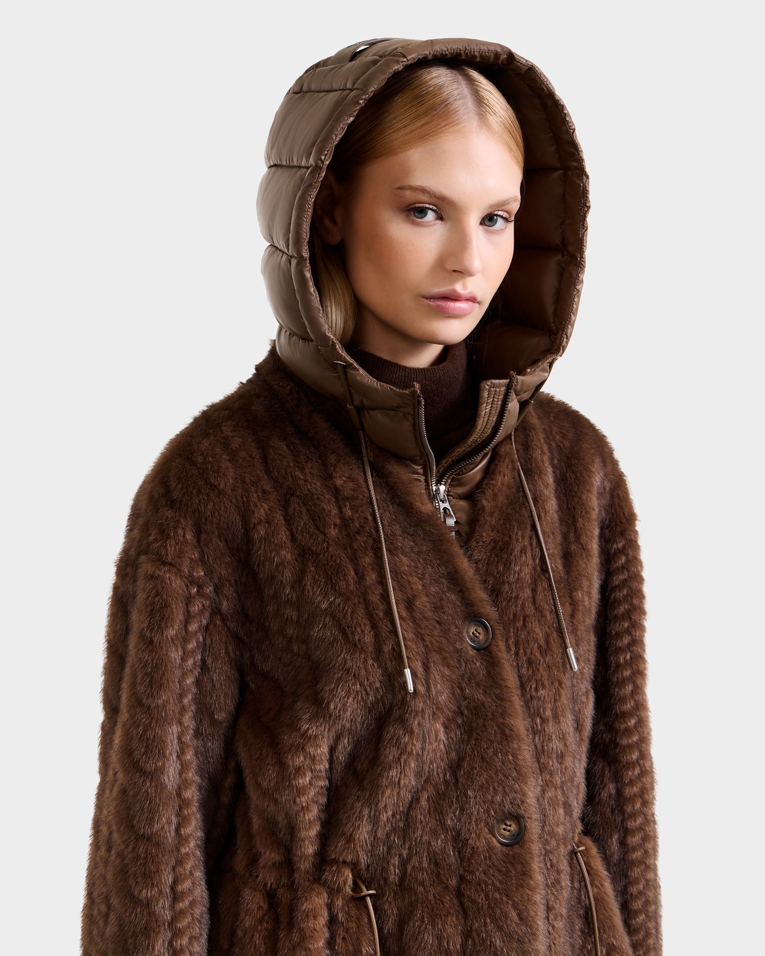 A woman with straight blonde hair models the Rudsak ORIANA - 8125545 Mocha women's faux fur coat with a shiny padded hood and vegan leather trim, standing against a plain light background with a neutral expression.
