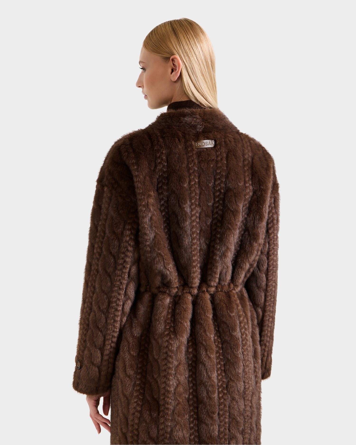 A person with straight blonde hair models the Rudsak ORIANA - 8125545 Mocha women’s long faux fur coat with vegan leather trim, shown from the back against a plain light gray background.