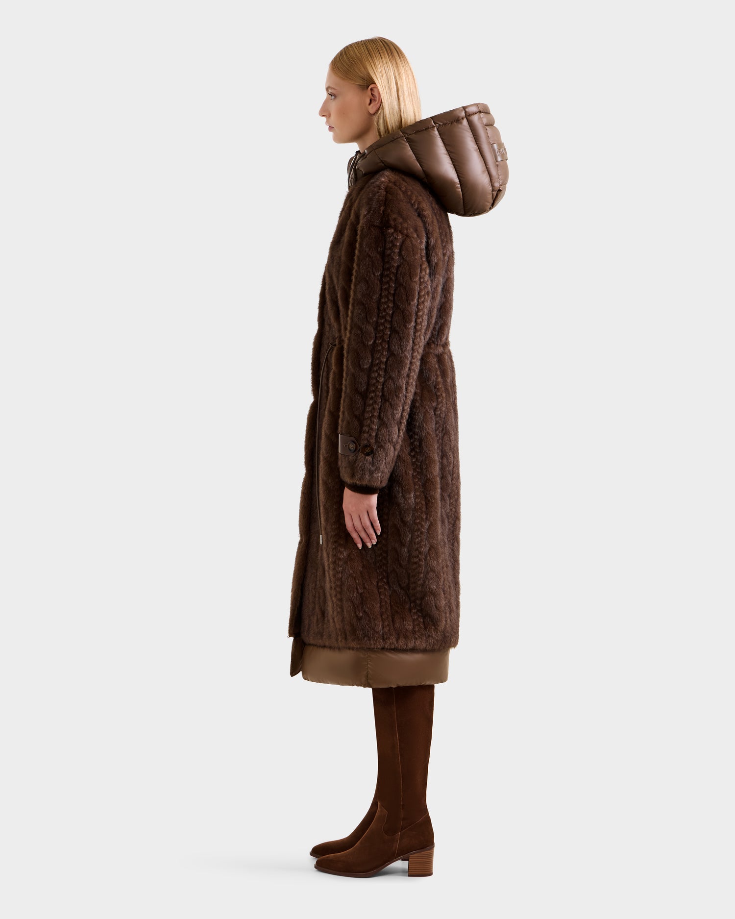 A woman in profile wears the Rudsak ORIANA - 8125545 Mocha faux fur coat with vegan leather trim and quilted hood, paired with brown knee-high boots and a beige dress, set against a plain light gray background.