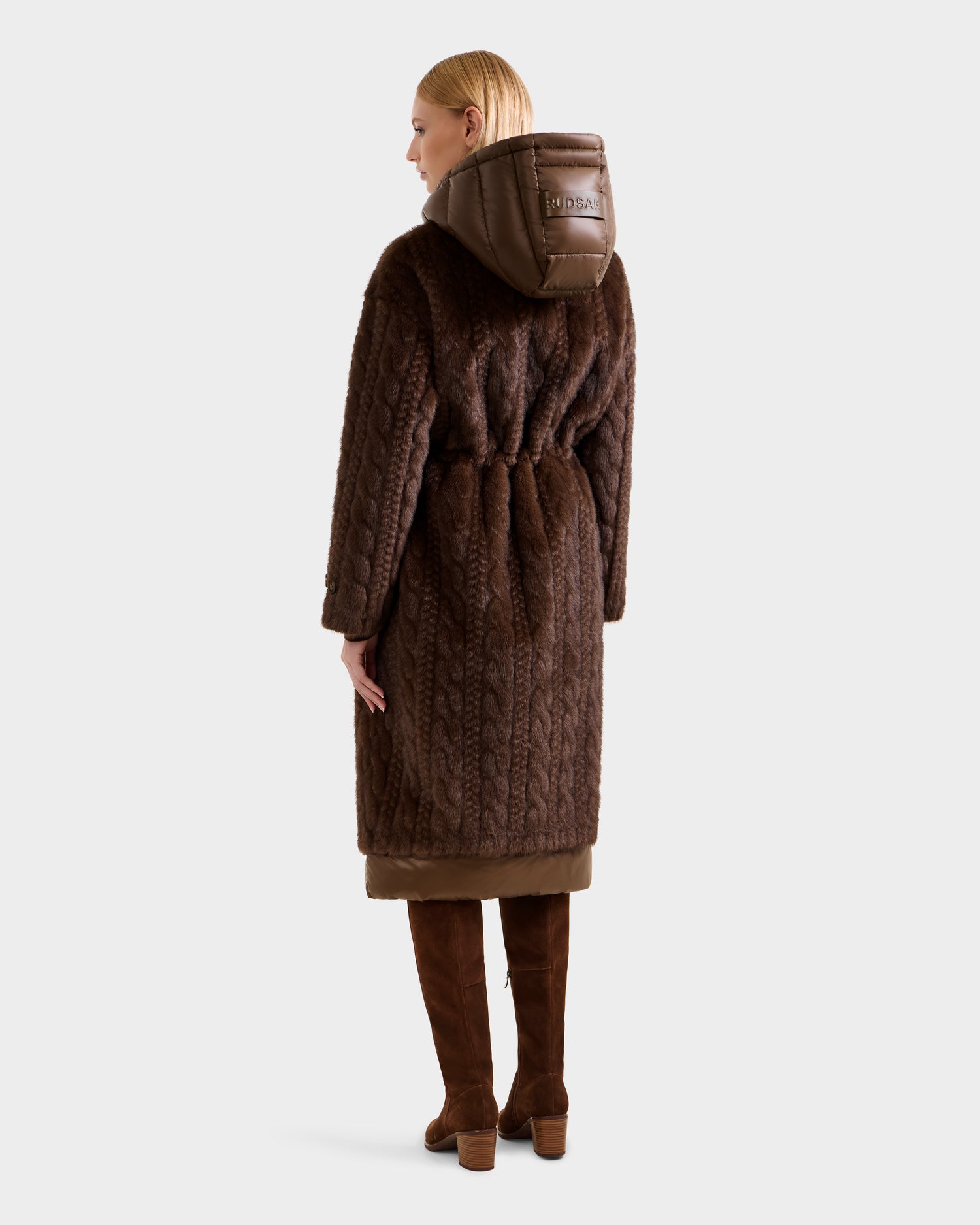 A person with blonde hair faces away, wearing the Rudsak ORIANA - 8125545 Mocha long dark brown cable knit hooded coat with a visible label and knee-high brown boots against a plain light background.