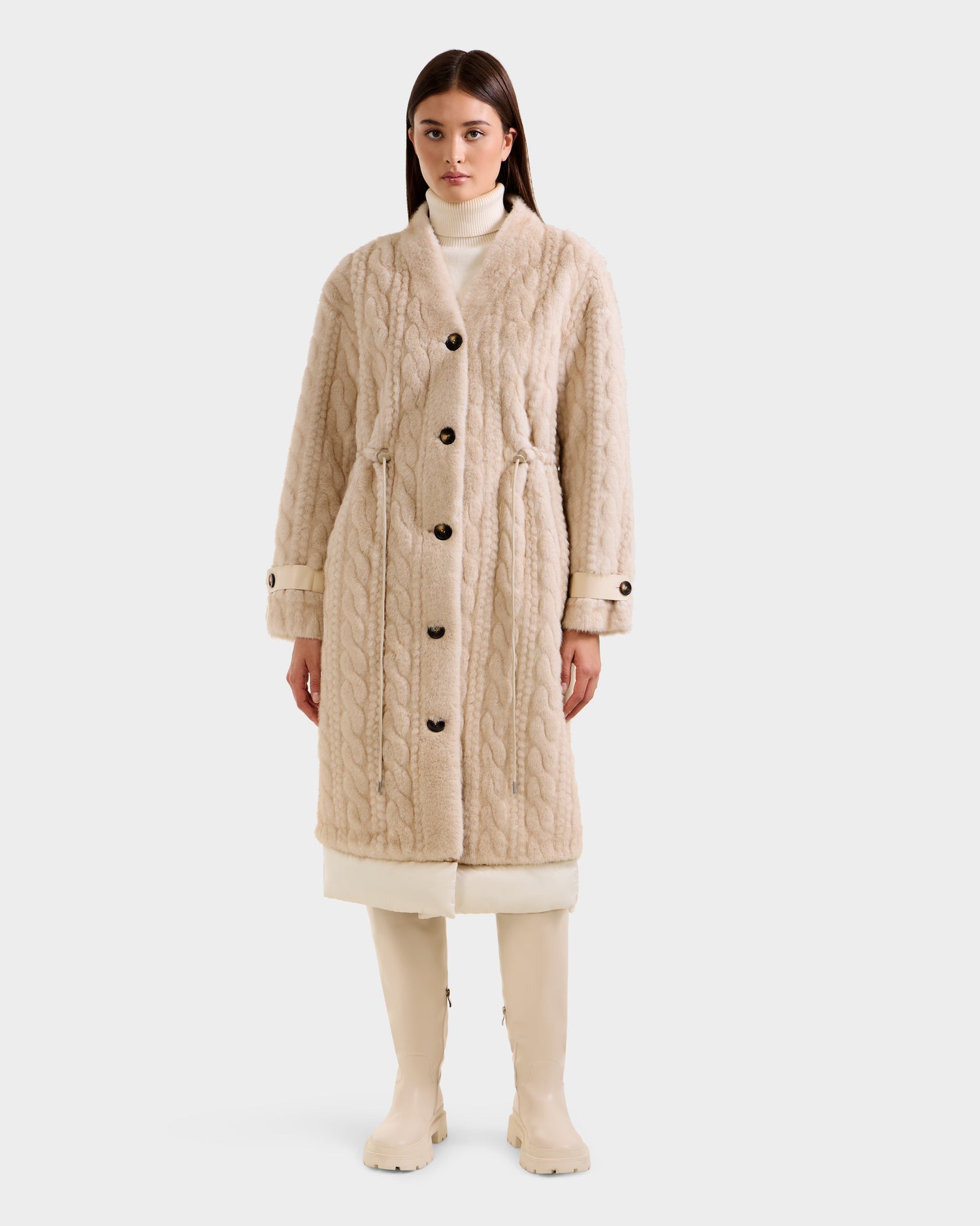 A woman stands facing forward in the Rudsak ORIANA - 8125545 Vanilla, a long beige cable knit coat with vegan leather trim and large buttons, styled with a white turtleneck and knee-high boots against a plain light background.
