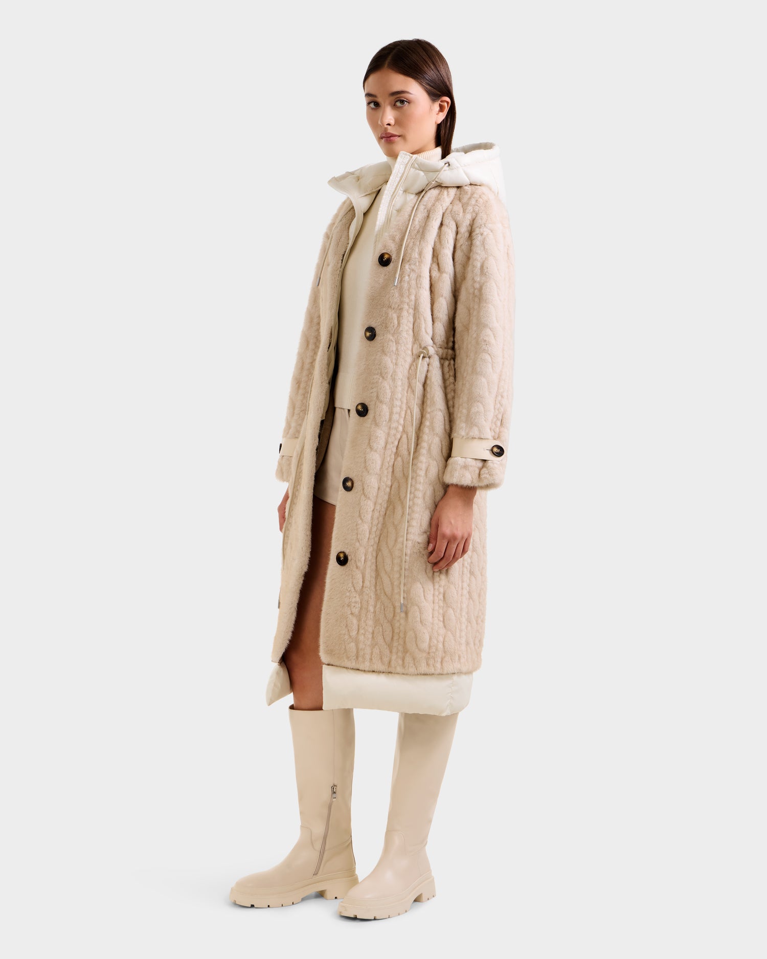 A woman stands against a white background wearing the Rudsak ORIANA - 8125545 Vanilla coat—knee-length, beige cable knit with buttons and vegan leather trim—paired with a high-collar cream sweater and knee-high cream boots.