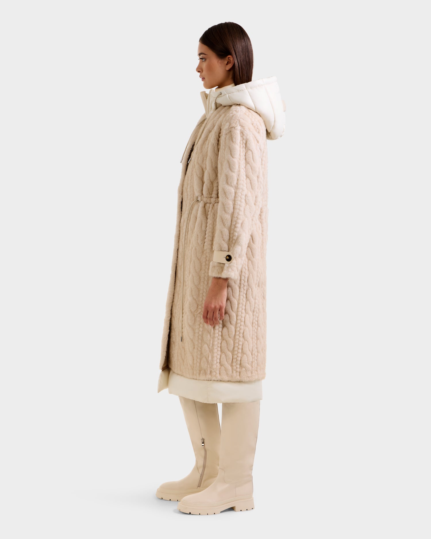 A woman in profile wears the Rudsak ORIANA - 8125545 Vanilla long beige cable knit hooded coat, a cream skirt or dress, and knee-high beige boots with vegan leather trim, set against a plain light background.