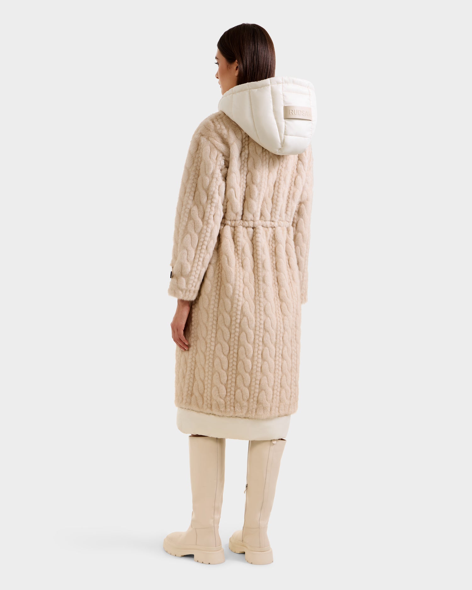 A woman faces away, wearing the Rudsak ORIANA - 8125545 Vanilla coat with a white hood, knee-high boots, vegan leather trim, belt detail at the waist, and textured pattern. The background is plain white.