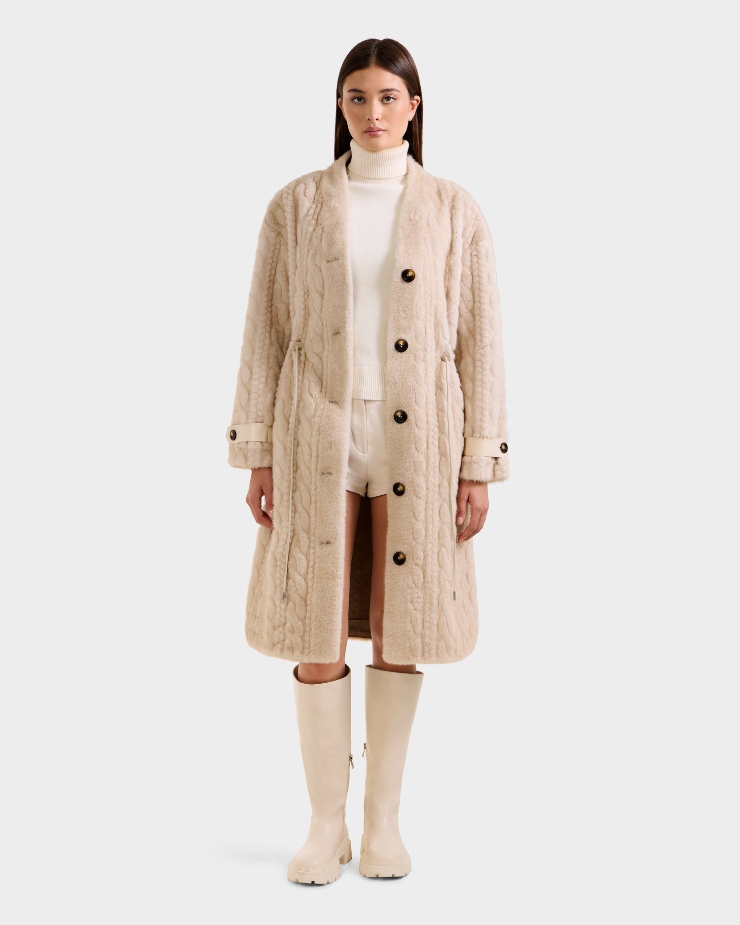 A woman faces forward in a Rudsak ORIANA - 8125545 Vanilla long beige cable knit coat with vegan leather trim and large buttons, layered over a white turtleneck and shorts, paired with knee-high cream boots, against a plain white background.