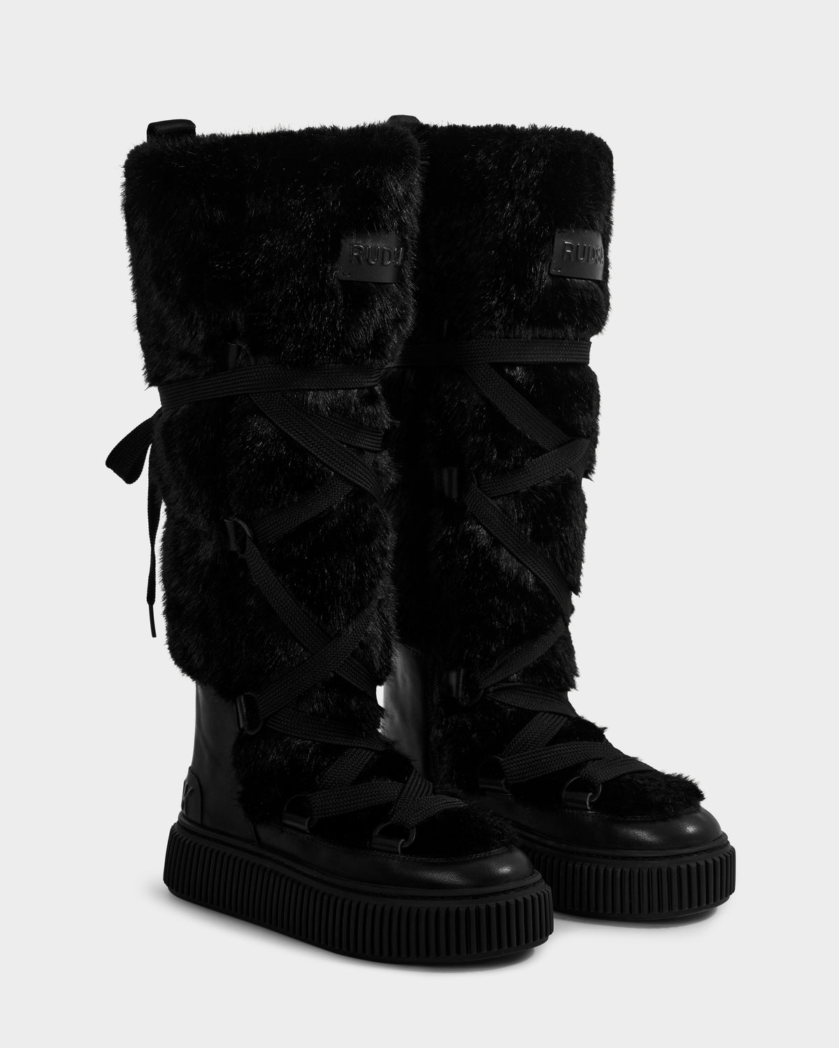 The Rudsak PALERMO C - 8225546 Black women's tall faux fur winter boots feature thick fur uppers, crisscrossing laces, rubber soles, and decorative side bows. Displayed on a white background—ideal après-ski style footwear.