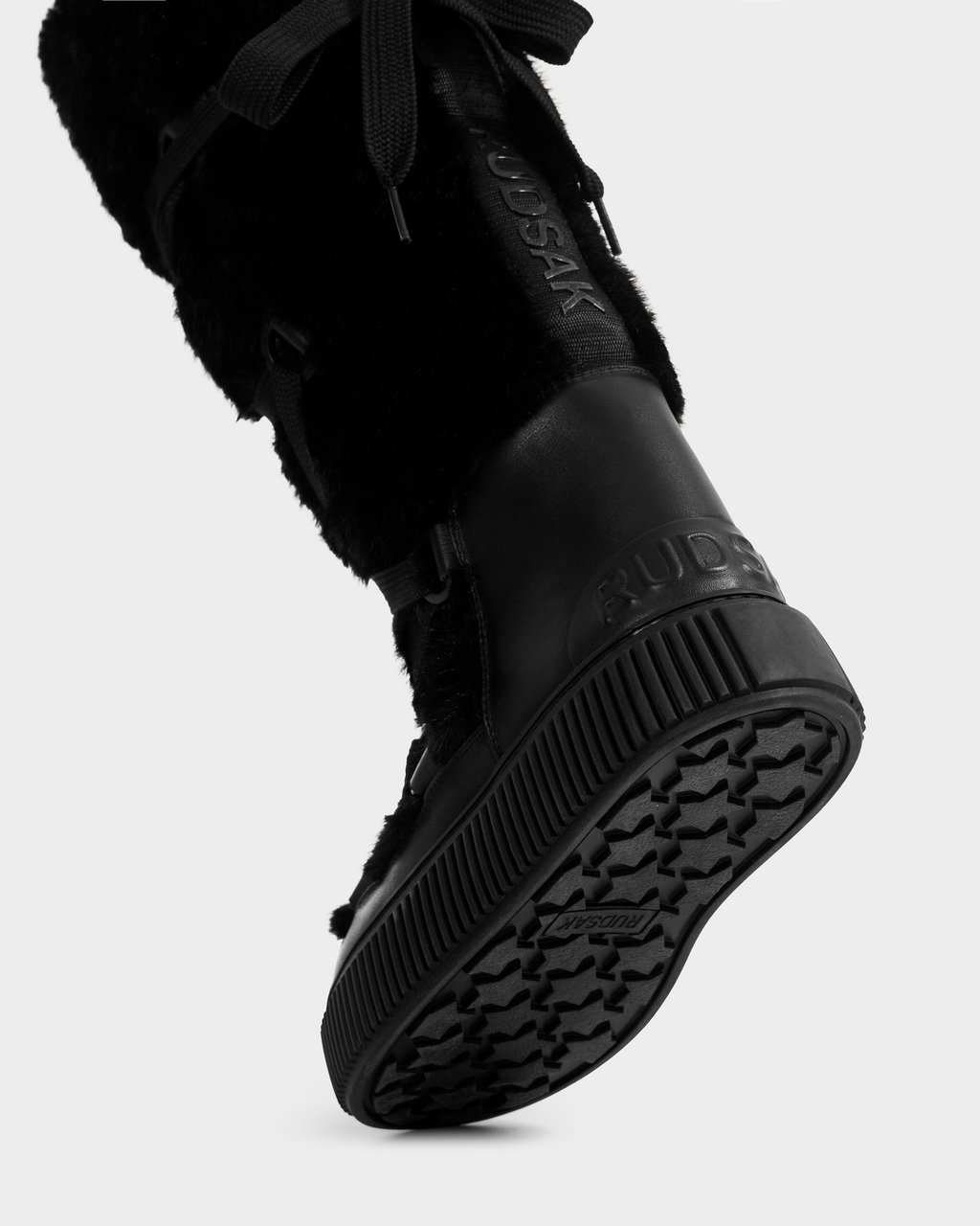 Close-up of a person’s foot wearing the Rudsak PALERMO C - 8225546 Black women’s winter boot with faux fur, textured sole, and brand details on the heel and side, standing out against a plain light gray background.