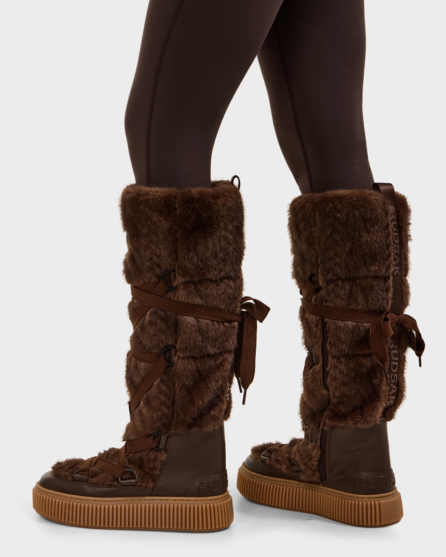 A person wearing dark brown leggings pairs them with Rudsak's PALERMO C - 8225546 Mocha boots—tall women’s faux fur winter boots featuring rubber soles and lace-up back details, reaching just below the knee.