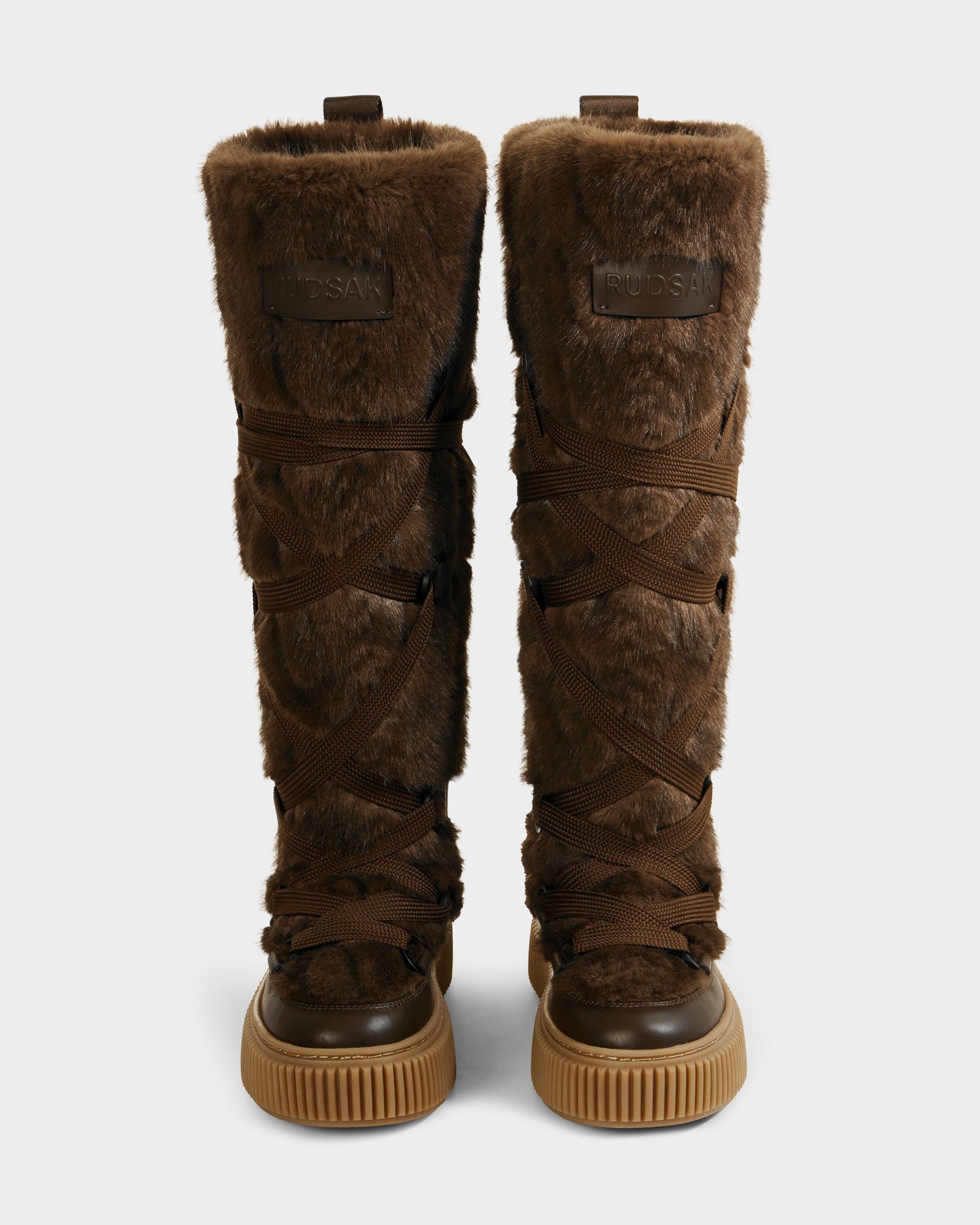 The Rudsak PALERMO C - 8225546 Mocha are tall women’s faux fur winter boots in brown with crisscross laces and tan rubber soles, shown upright on a light gray background—ideal for cozy après-ski and winter style.