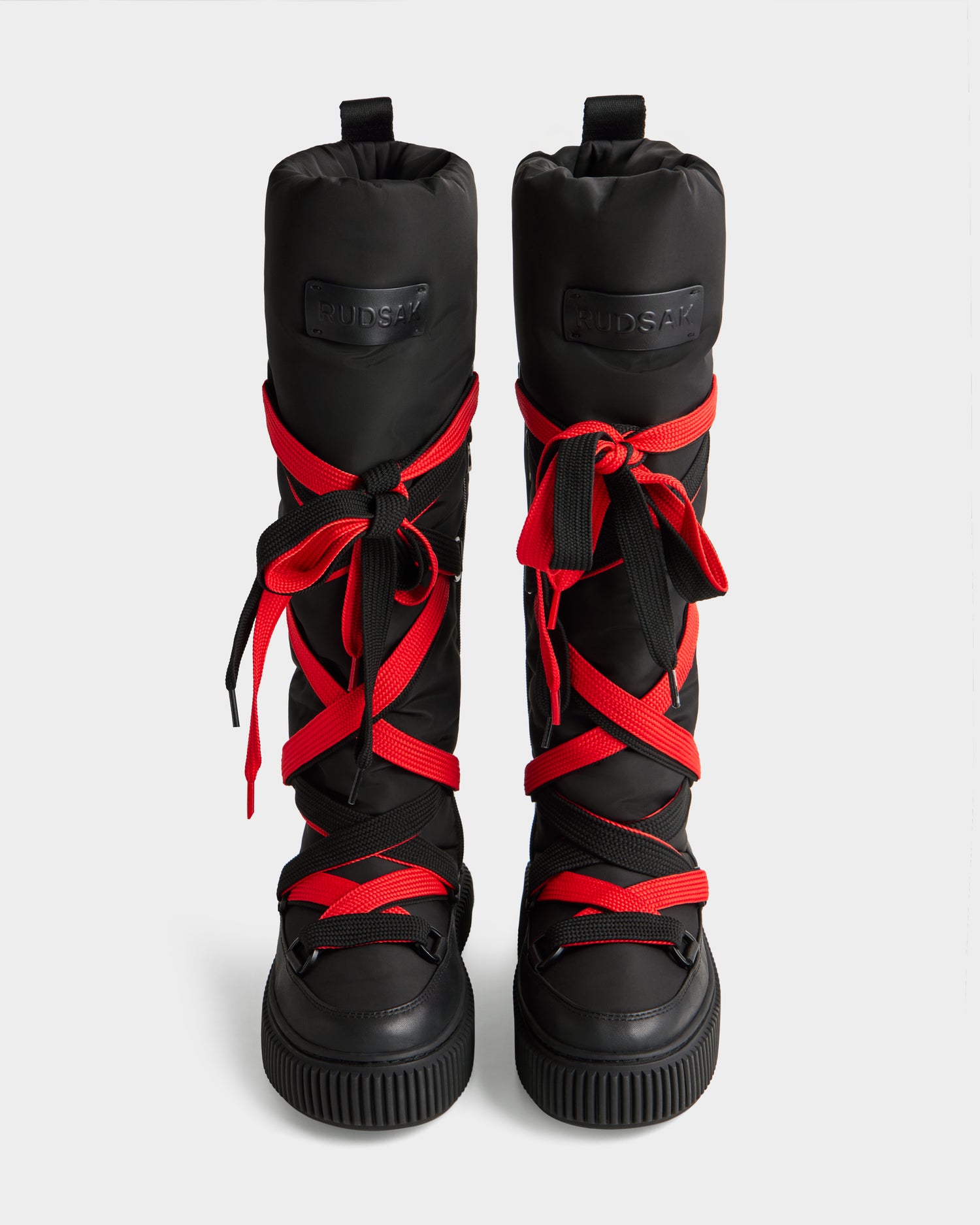 The Rudsak PALERMO - 8225030 Black boots feature tall, insulated uppers, ribbed soles, and thick red-and-black crisscross laces, shown from the front against a plain light background.