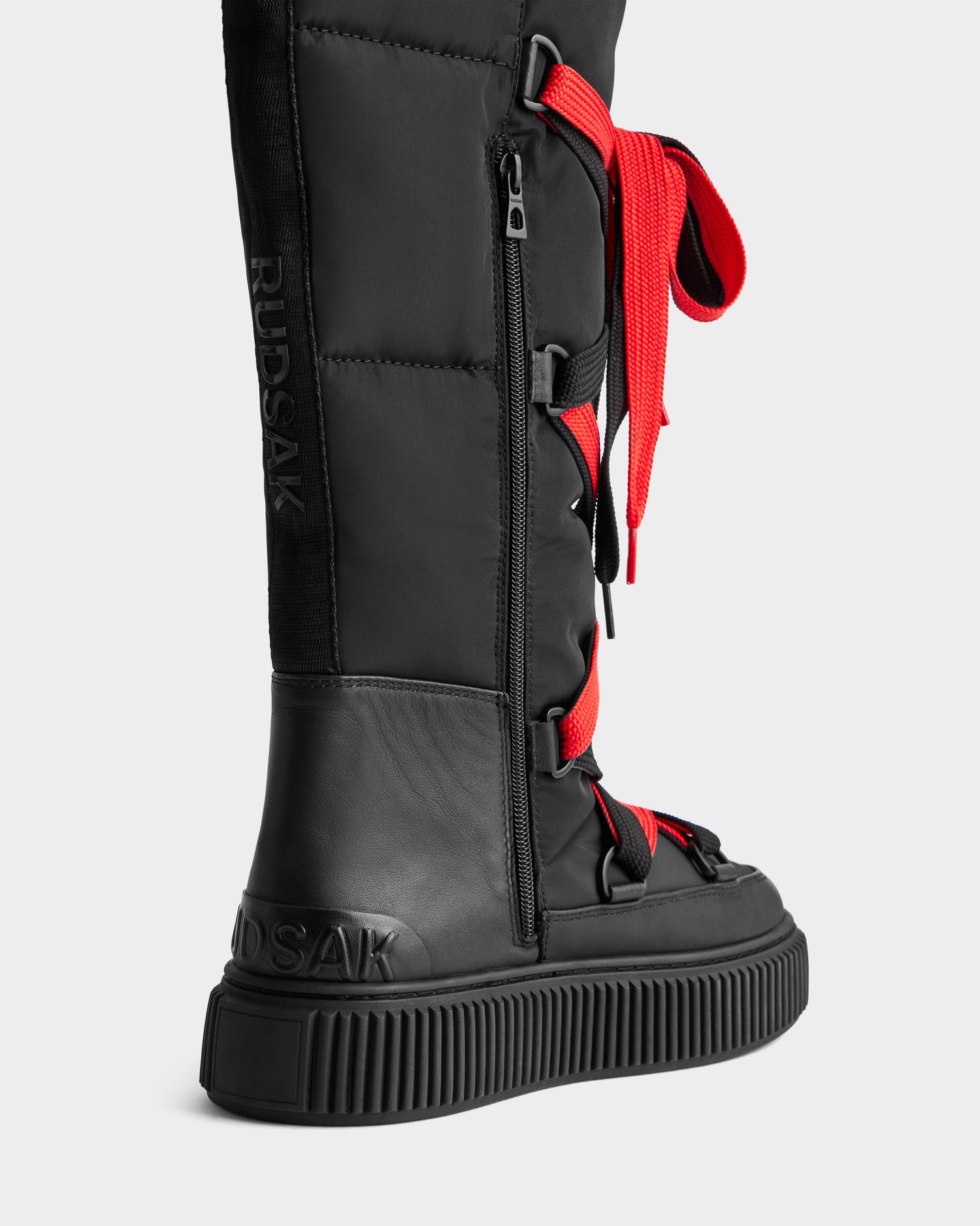 A close-up of the Rudsak PALERMO - 8225030 Black women's knee-high winter boot, featuring thick textured sole, red laces, side zipper, and embossed branding for a sporty yet functional modern design.