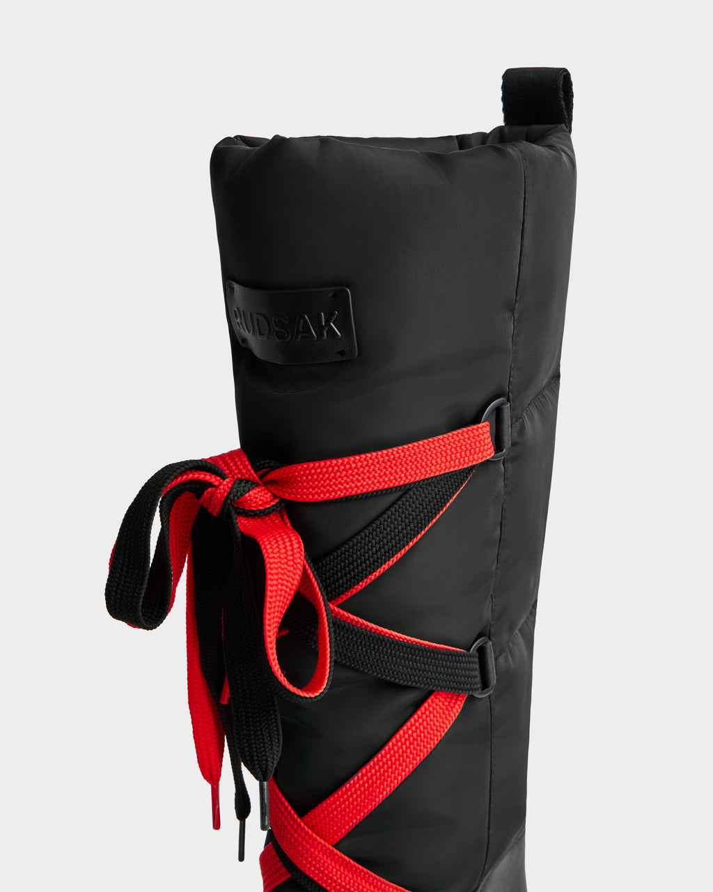 A close-up of the Rudsak PALERMO - 8225030 Black women’s winter boot with red and black laces, a black pull tab, padded insulated exterior, and a branded patch.