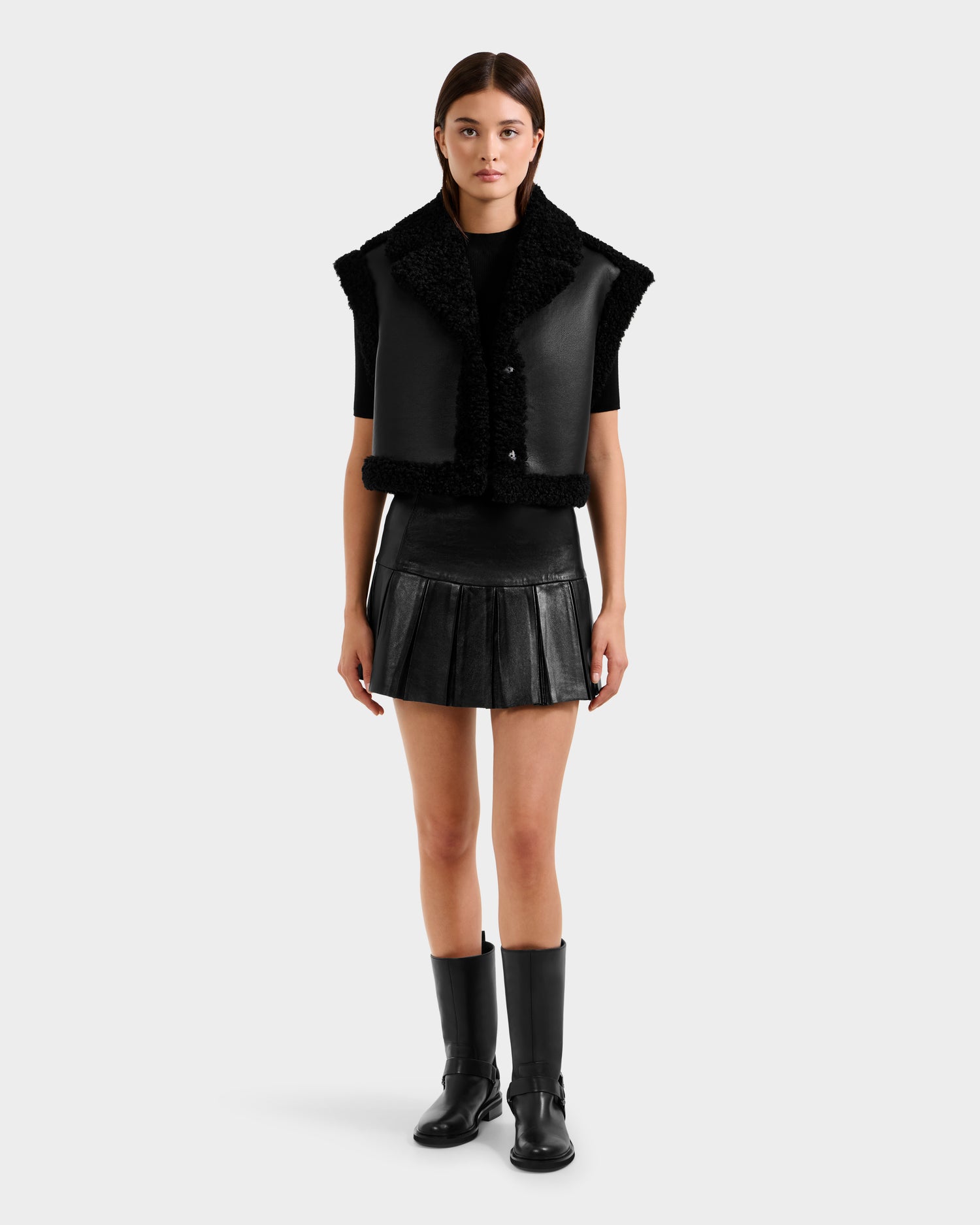 A woman faces forward against a white background, wearing the Rudsak PALOMA - 8125675 Black faux shearling short-sleeve vest, a black pleated mini skirt, black t-shirt, and knee-high black boots.