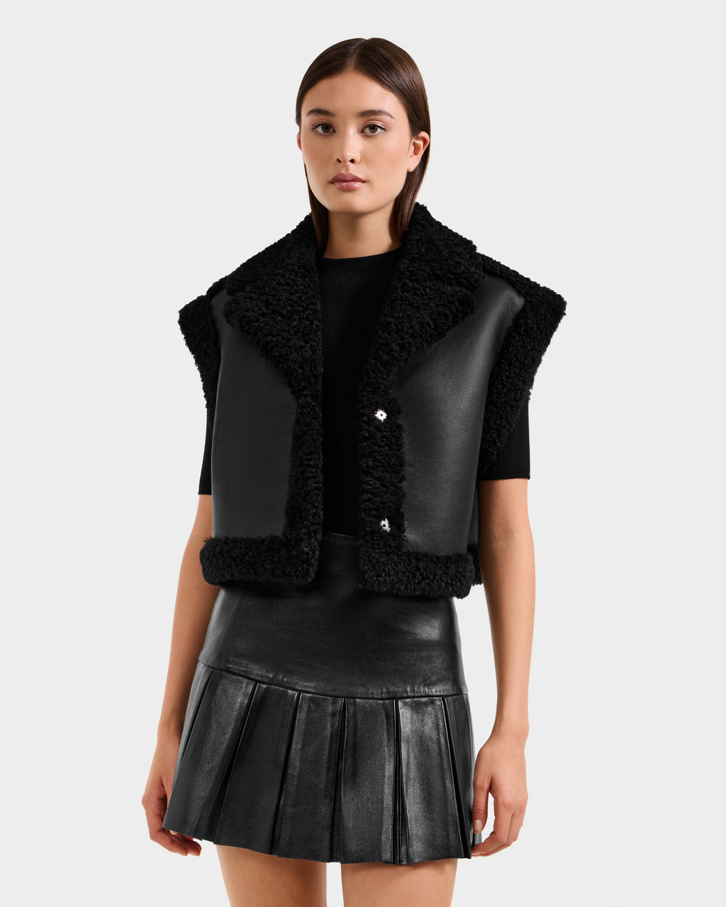 A woman wears the Rudsak PALOMA - 8125675 Black faux shearling vest over a black short-sleeve top and pleated leather skirt, standing against a light gray background.