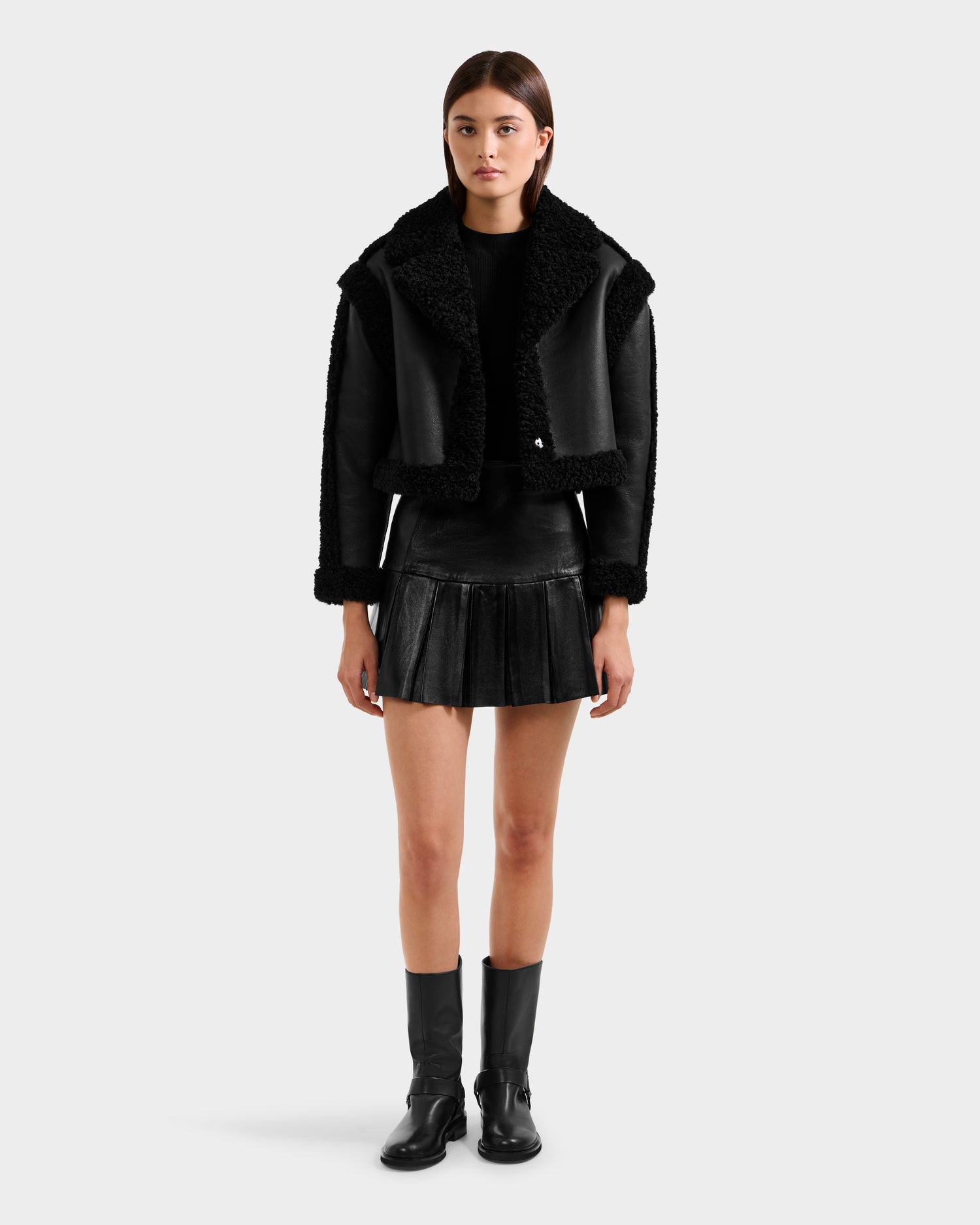 A woman with straight hair and a neutral expression stands against a plain background wearing the Rudsak PALOMA - 8125675 Black faux shearling jacket, paired with a black pleated mini skirt, black top, and mid-calf boots.