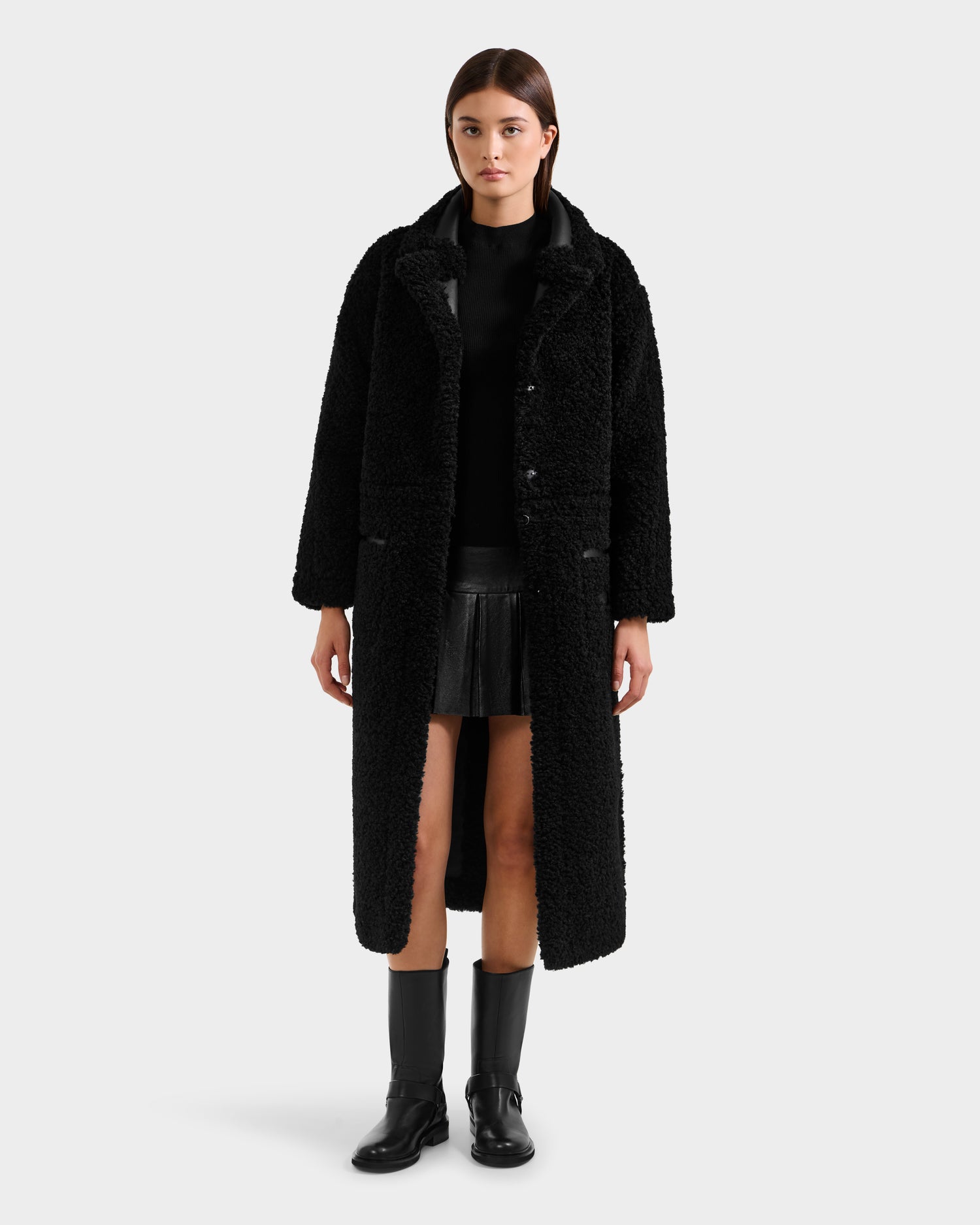 A woman faces forward in a Rudsak PALOMA - 8125675 Black faux shearling coat, paired with a black top, pleated skirt, and boots, set against a plain light gray background.
