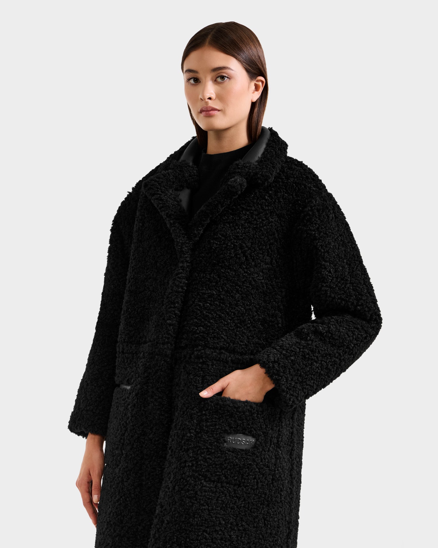 A person with straight brown hair models the Rudsak PALOMA - 8125675 Black faux shearling coat, featuring a wide collar and large front pockets, against a plain light gray background. One hand rests in a pocket; their expression is neutral.