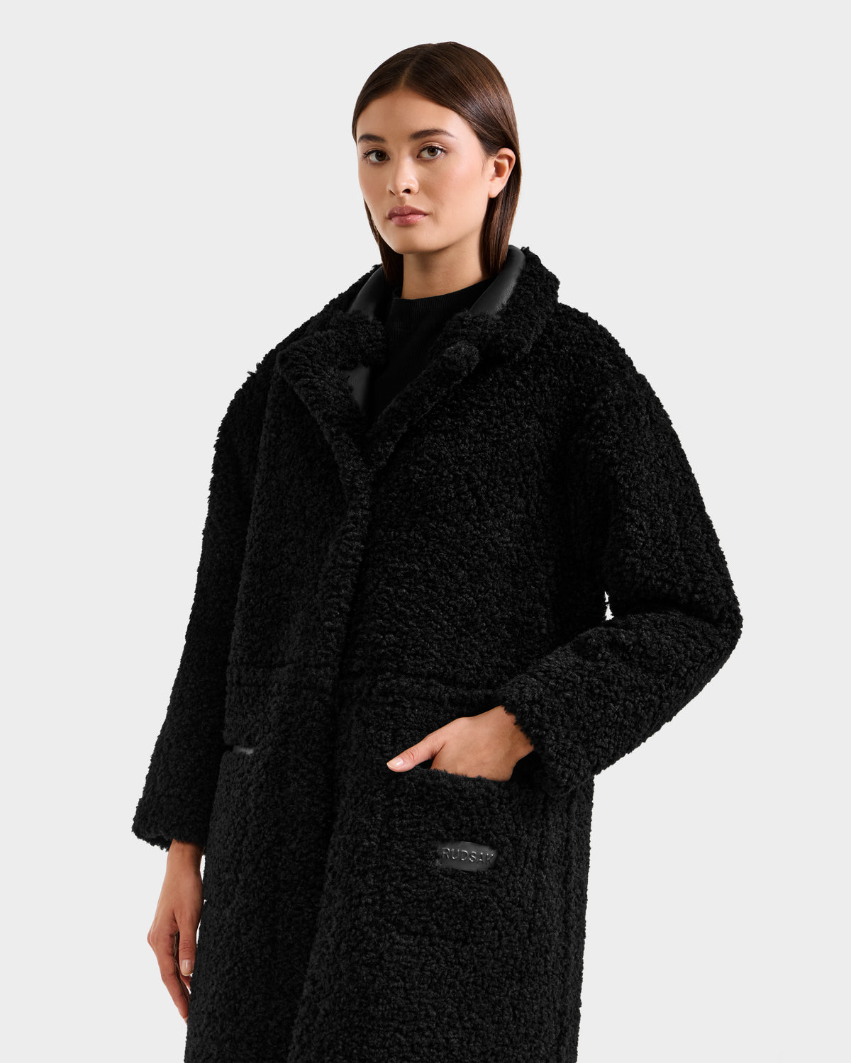 A person with straight brown hair models the Rudsak PALOMA - 8125675 Black faux shearling coat, featuring a wide collar and large front pockets, against a plain light gray background. One hand rests in a pocket; their expression is neutral.