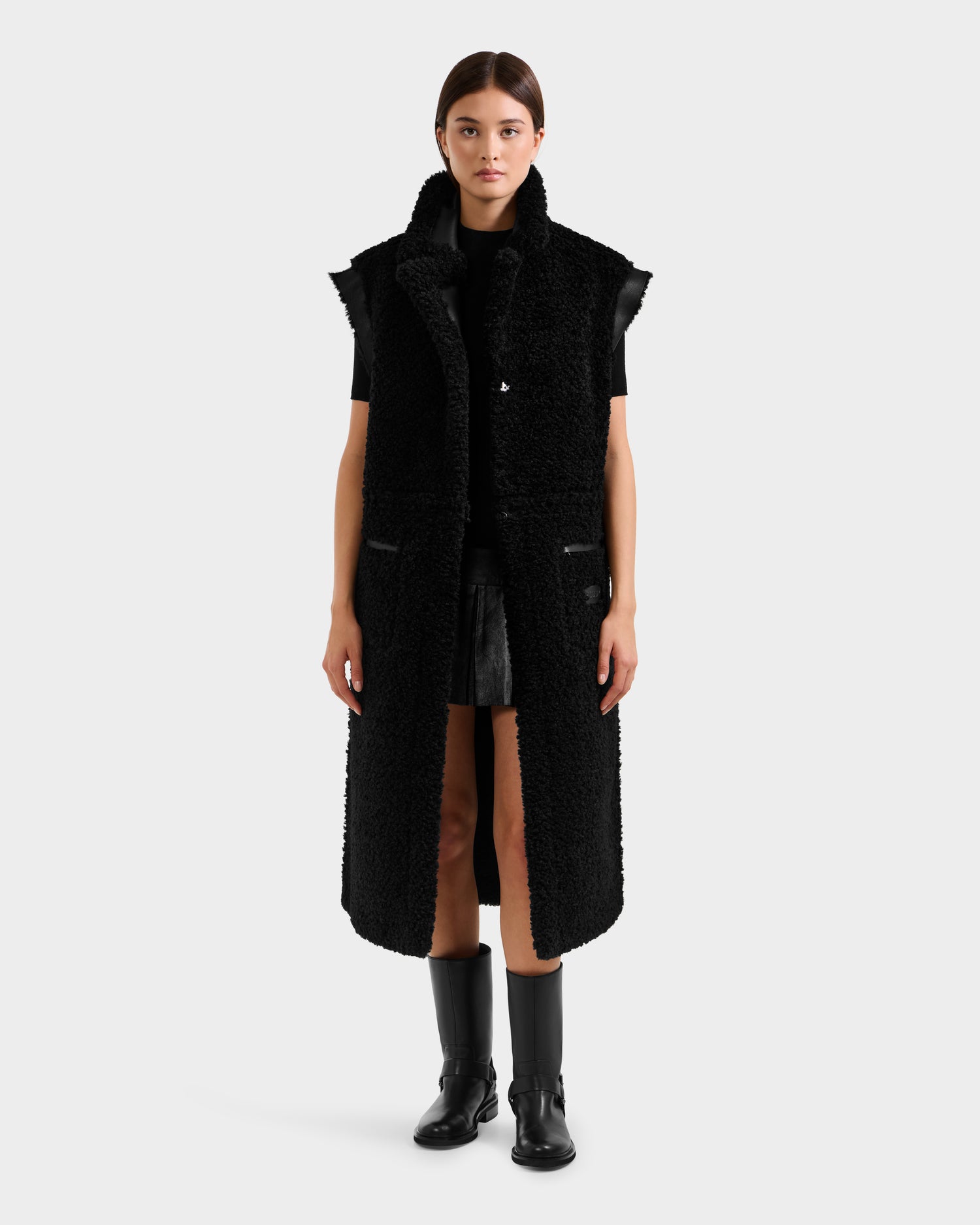 A woman faces forward in a Rudsak PALOMA - 8125675 Black sleeveless faux shearling long vest over a black outfit and knee-high boots, highlighting chic modular outerwear against a plain light gray background.