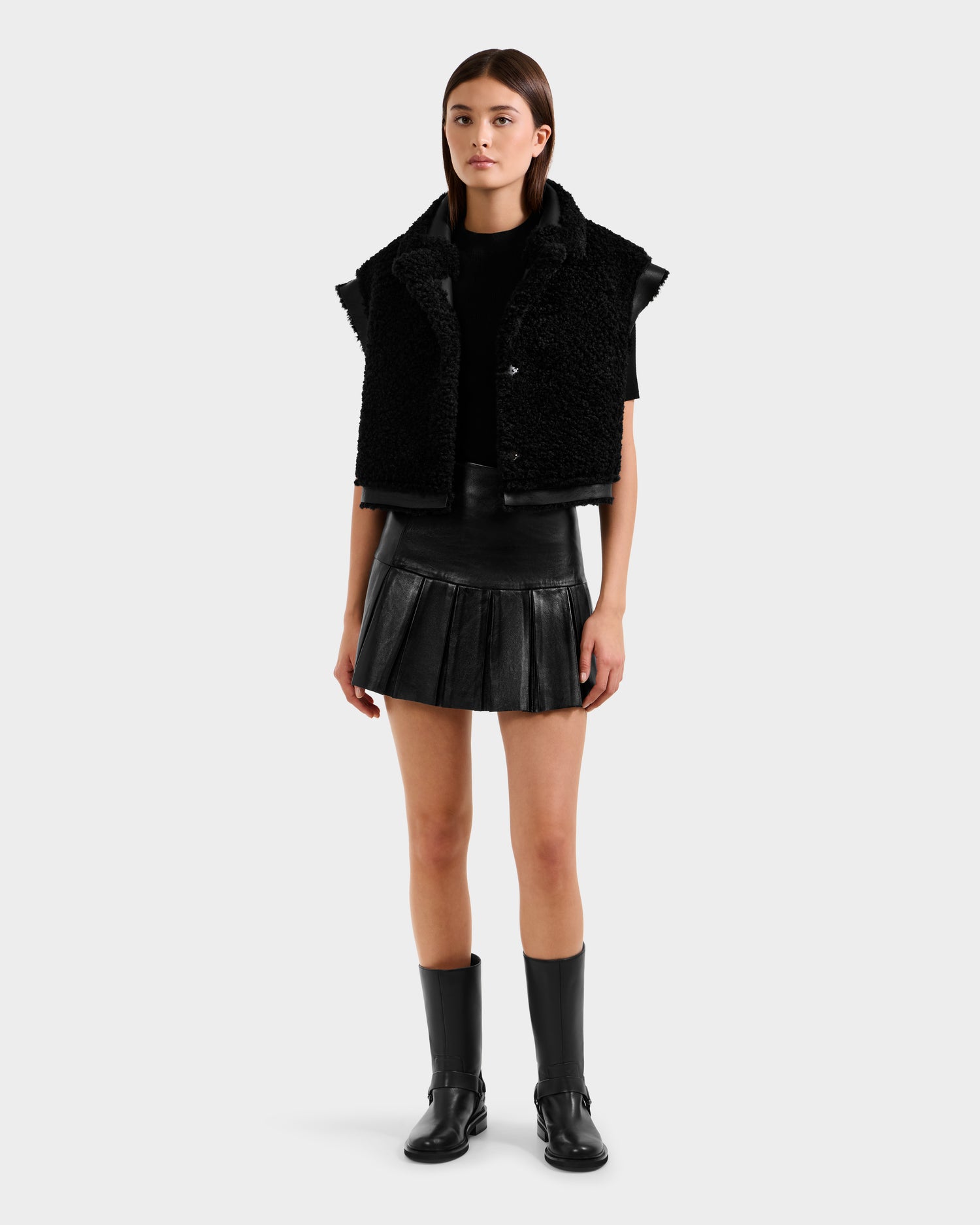 A woman with straight brown hair and a neutral expression wears the Rudsak PALOMA - 8125675 Black modular outerwear: a black textured vest over a black top, pleated leather mini skirt, and black boots against a plain background.
