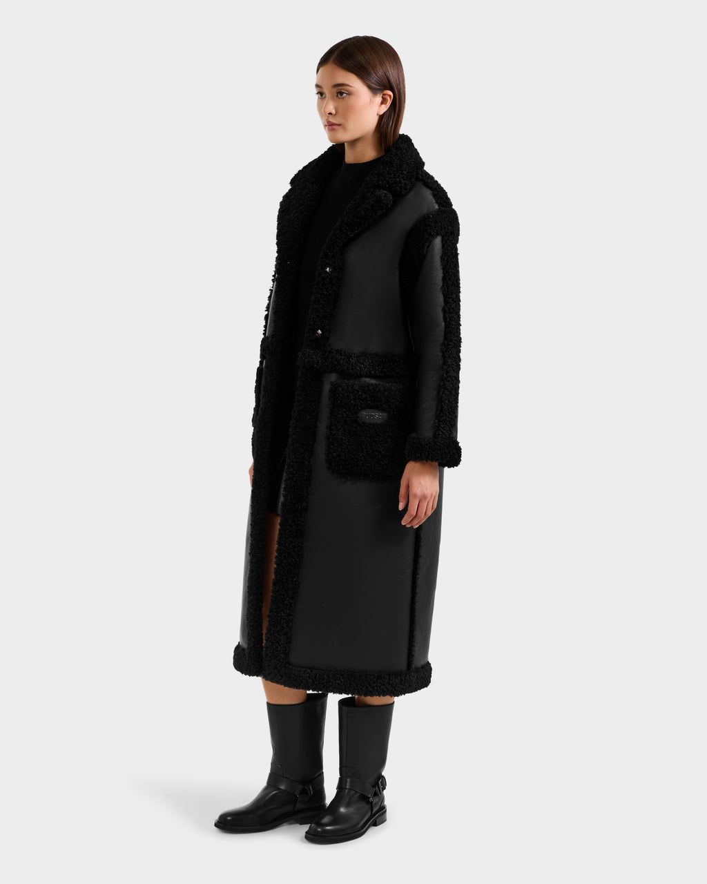 A woman models the Rudsak PALOMA - 8125675 Black faux shearling coat with textured trim, paired with a black top and knee-high boots, standing slightly turned against a plain light gray background.